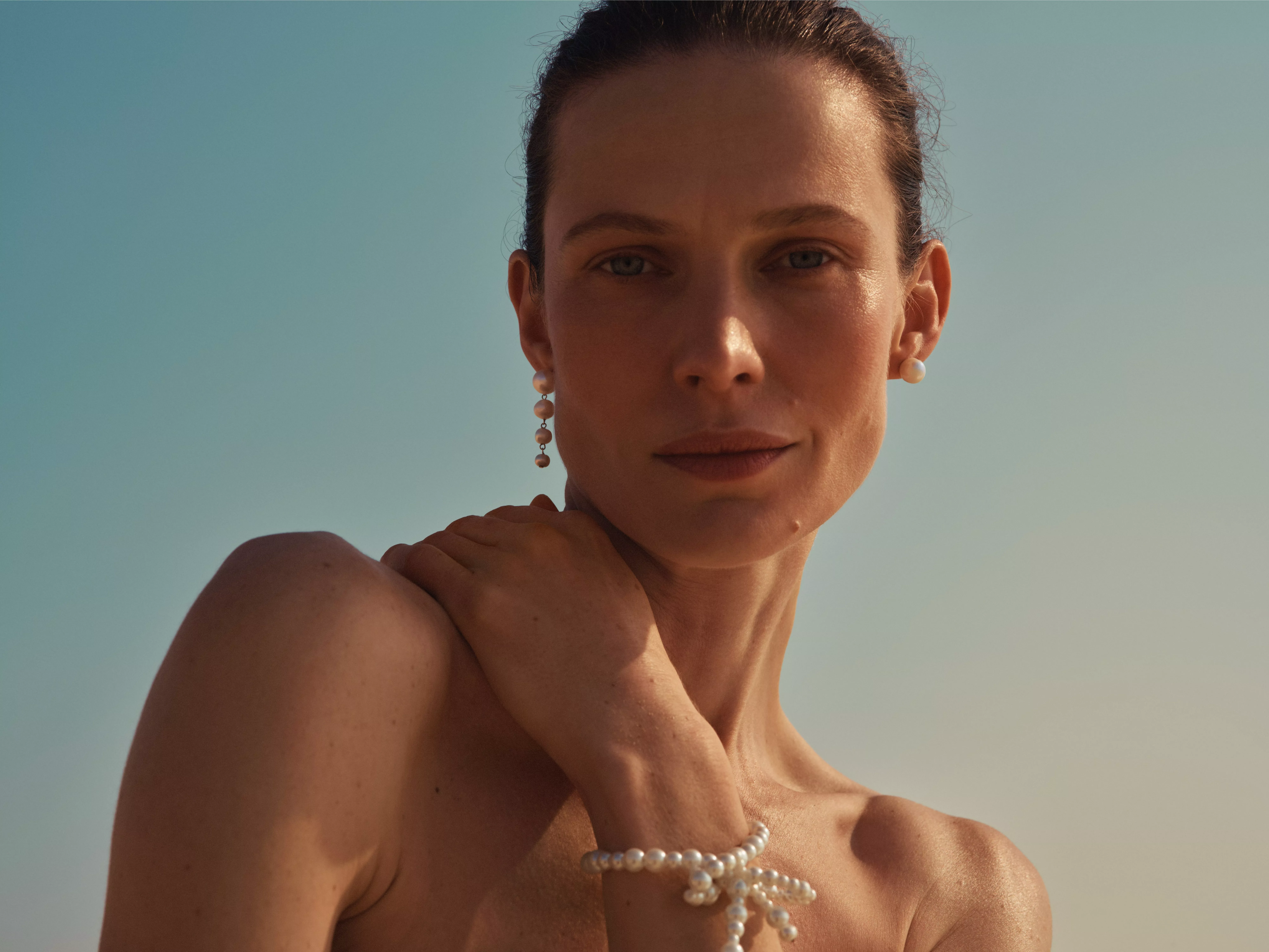 Close-up portrait of a woman wearing pearl earrings and bracelet, posing outdoors against a clear blue sky