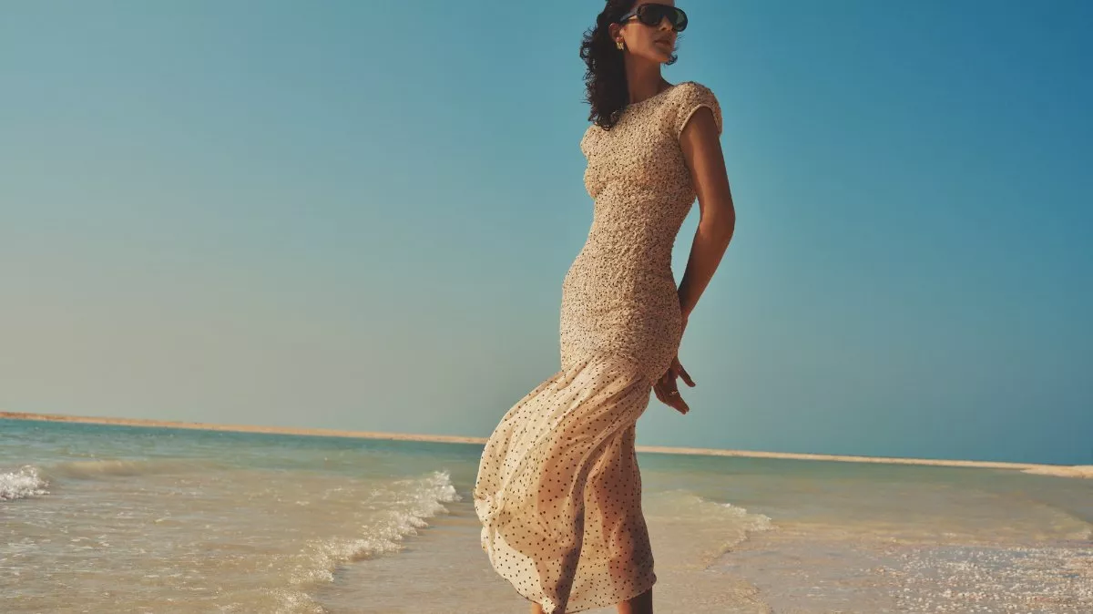 Woman in a beige maxi dress and sunglasses standing by the sea, luxury summer fashion photoshoot at sunset