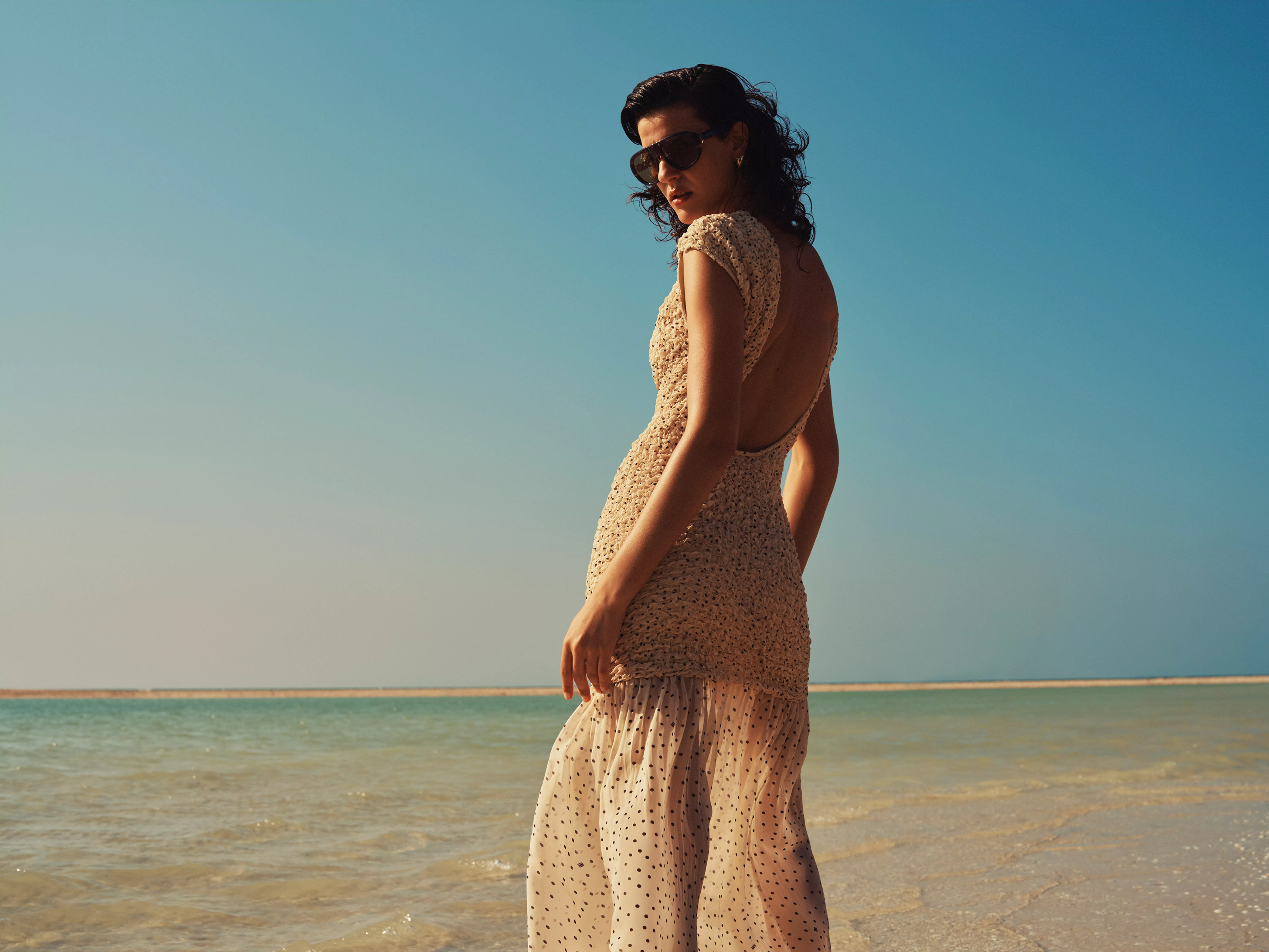 Woman in sunglasses wearing a beige crochet open-back dress standing by the sea under a clear blue sky