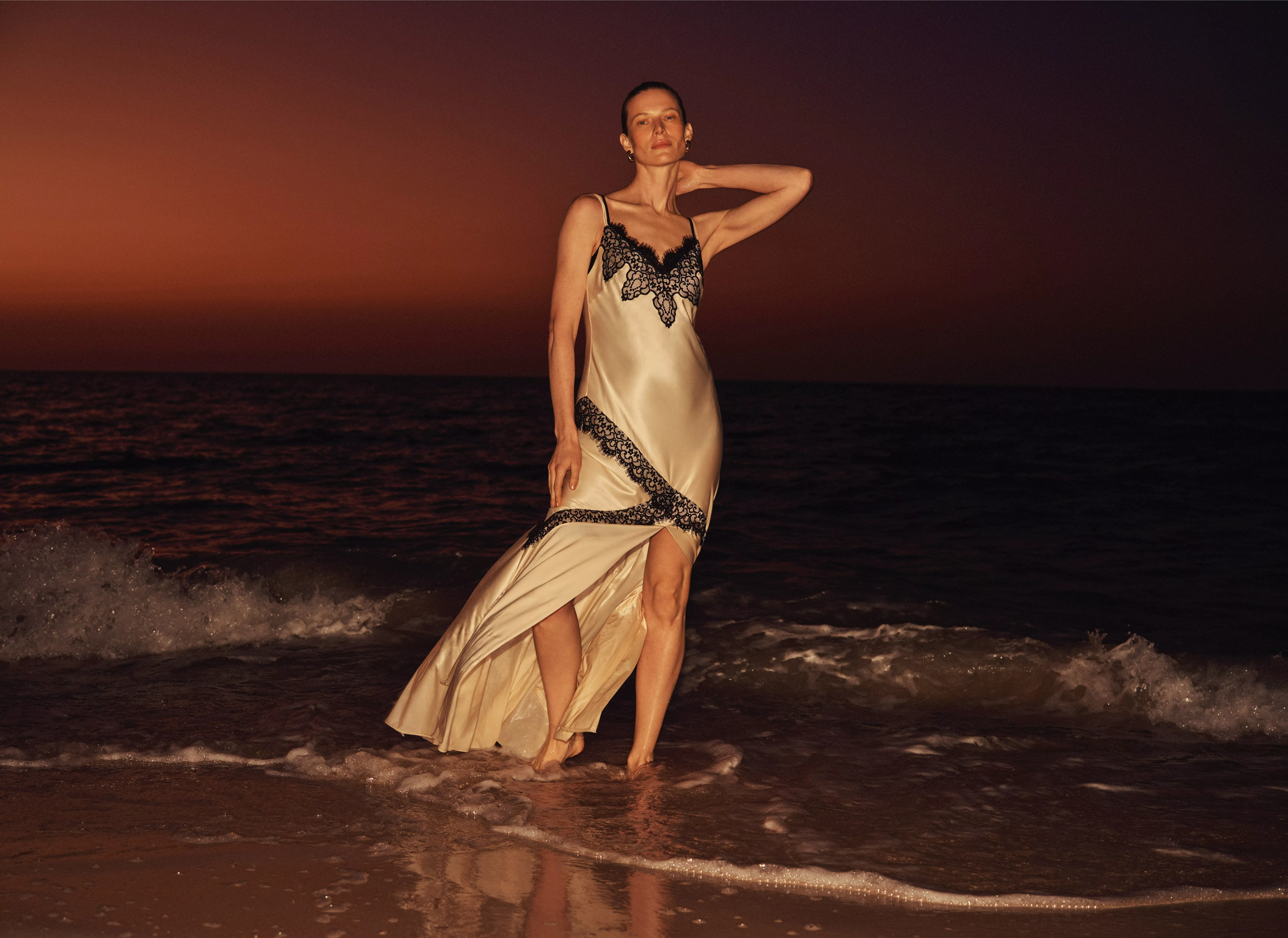 A person in a flowing silk dress with black lace details, stands in shallow sea waves at sunset, with a vibrant orange sky.