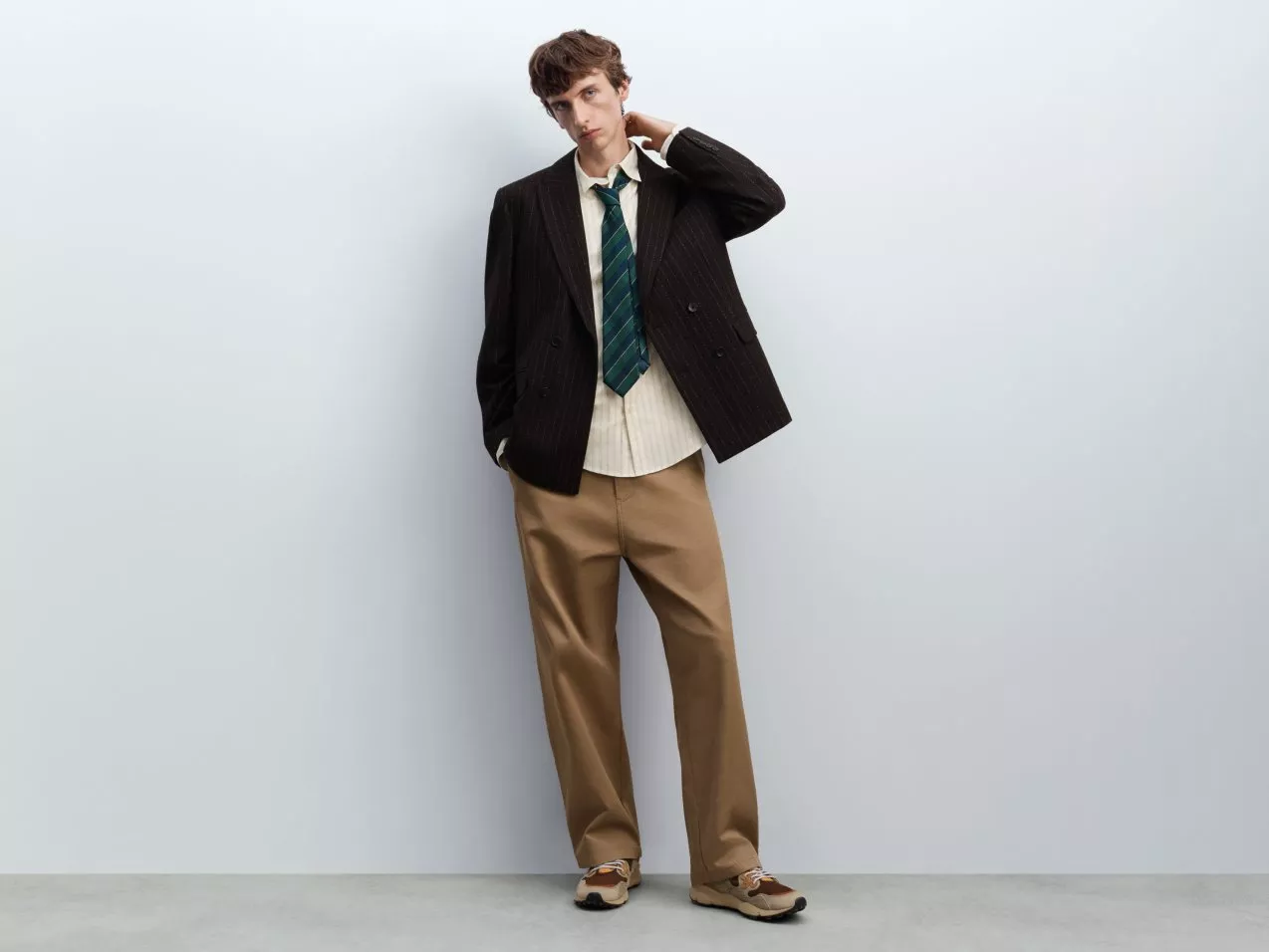 Full-length fashion portrait of a man in pinstripe blazer, striped tie and tan trousers against grey wall