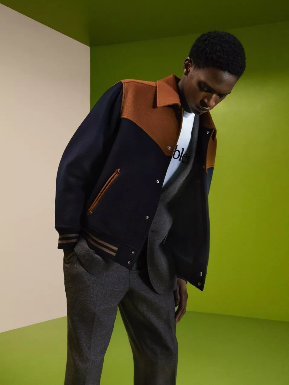 Male fashion model wearing navy and tan varsity jacket with grey suit trousers in a green studio backdrop