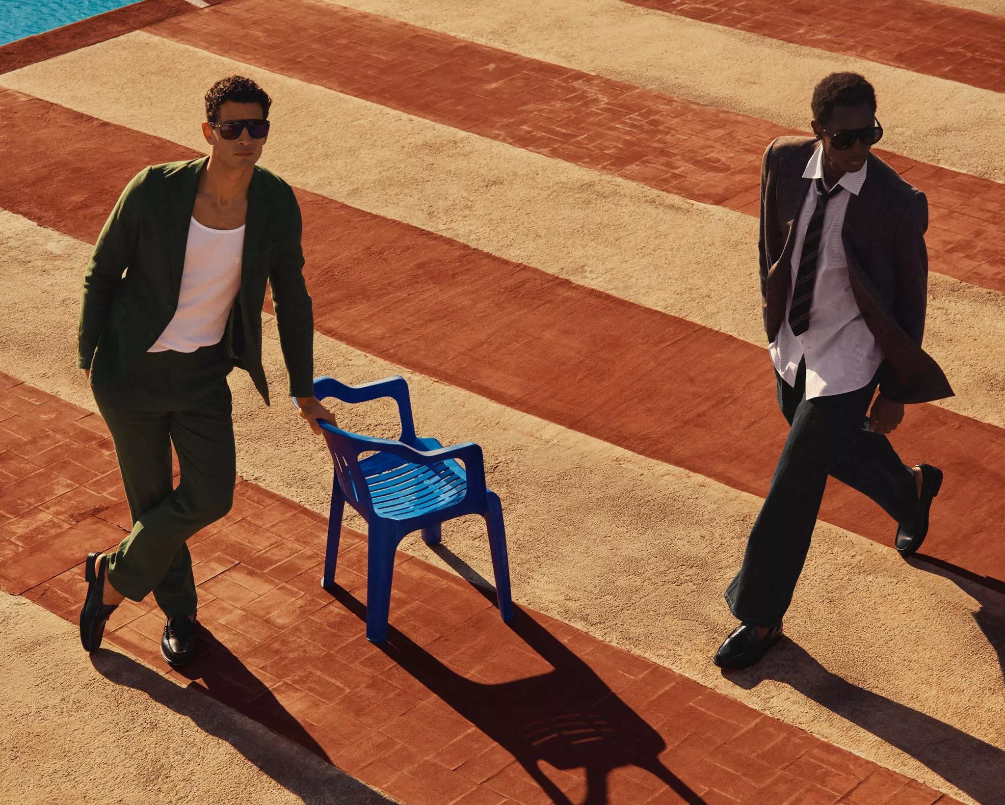 Two stylish men in tailored suits and sunglasses walking by a pool on striped tiles, luxury menswear fashion shoot