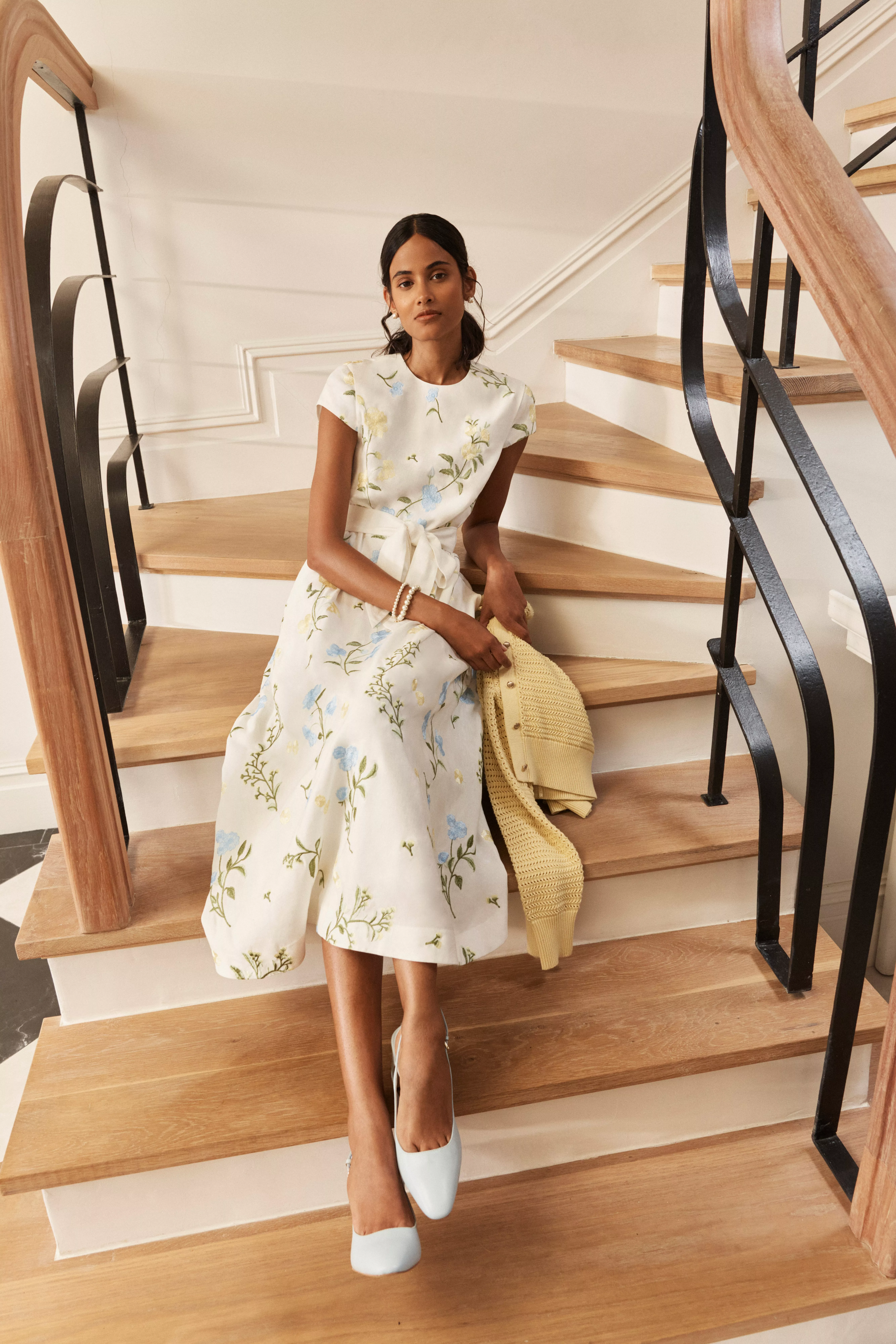 Woman sitting on wooden staircase in floral midi dress and blue heels, holding a yellow knit cardigan indoors