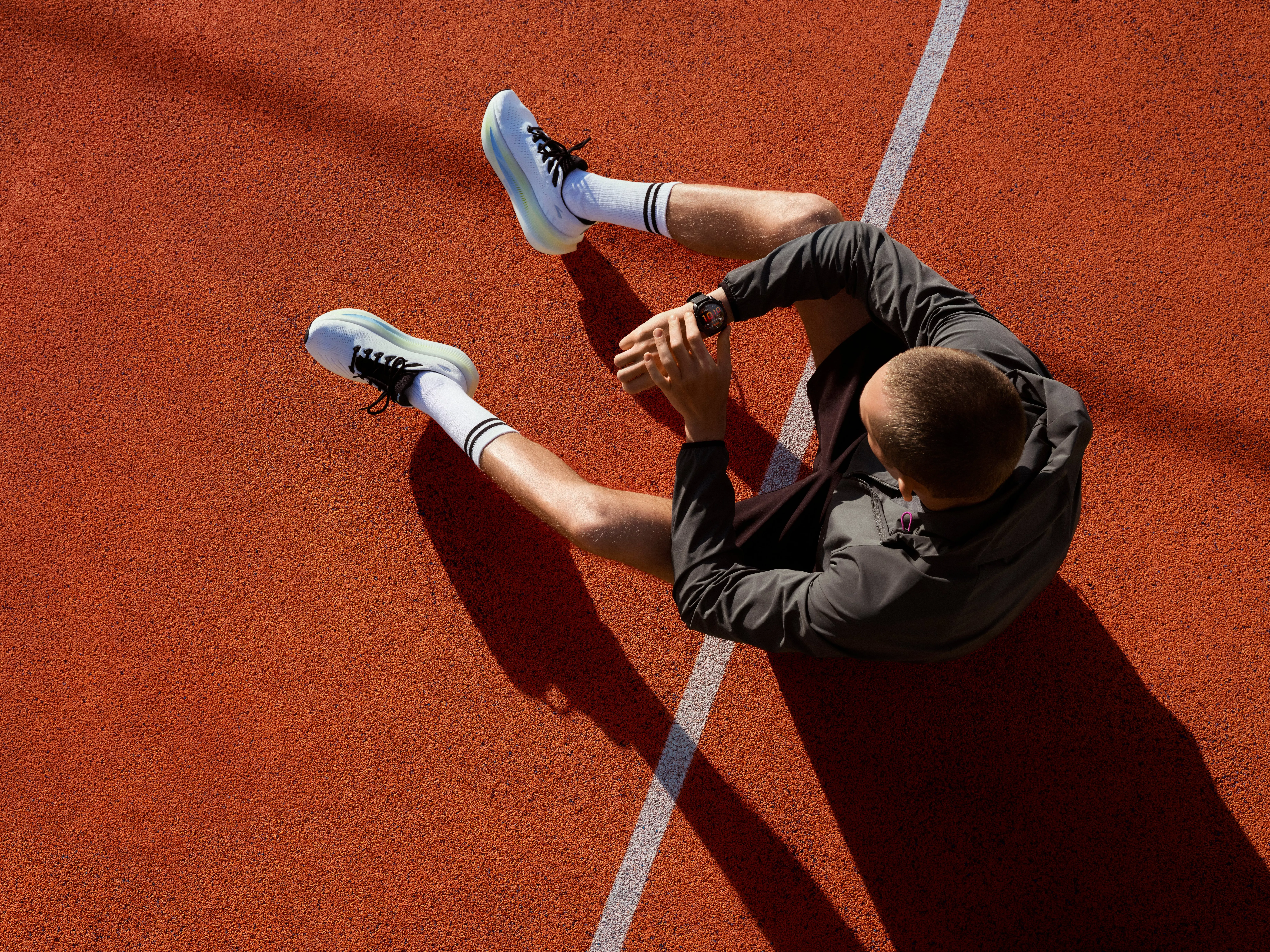 Overhead view of athlete stretching on a running track, wearing trainers, socks and sports jacket in sunlight
