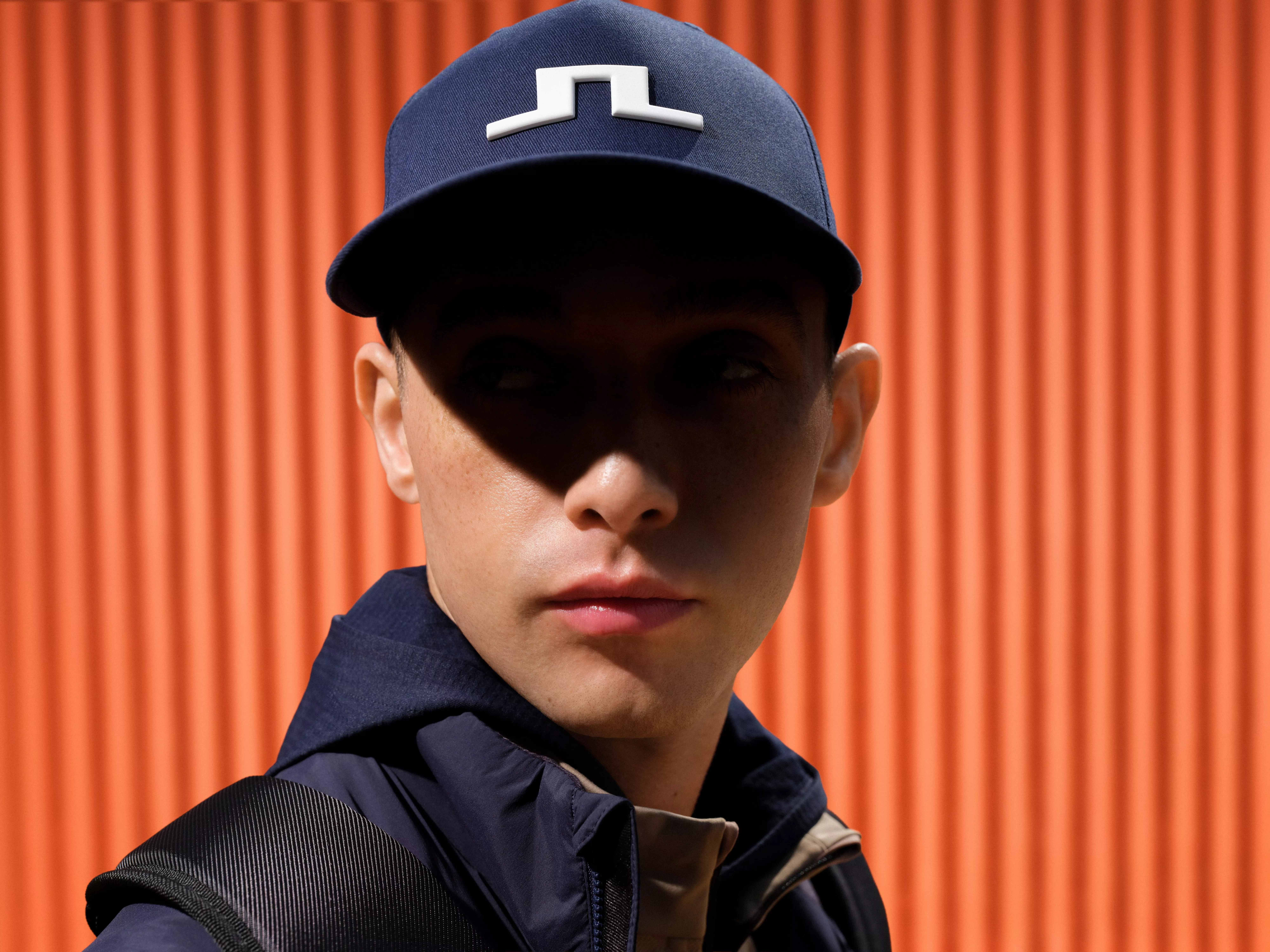Close-up portrait of man in navy baseball cap and jacket with backpack, against orange corrugated wall backdrop