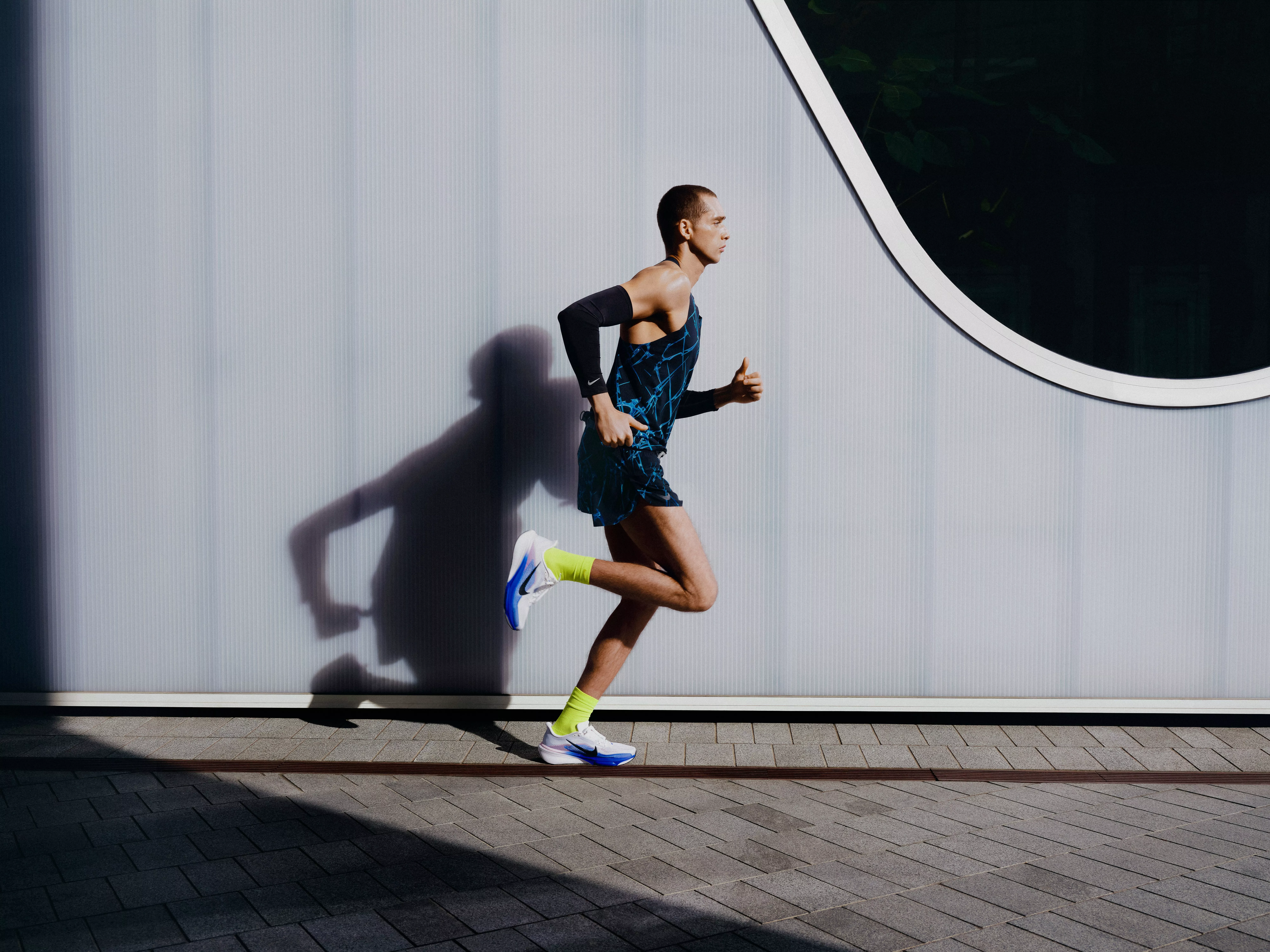 Male runner sprinting outdoors beside a modern building wall, wearing performance trainers and sportswear in sunlight
