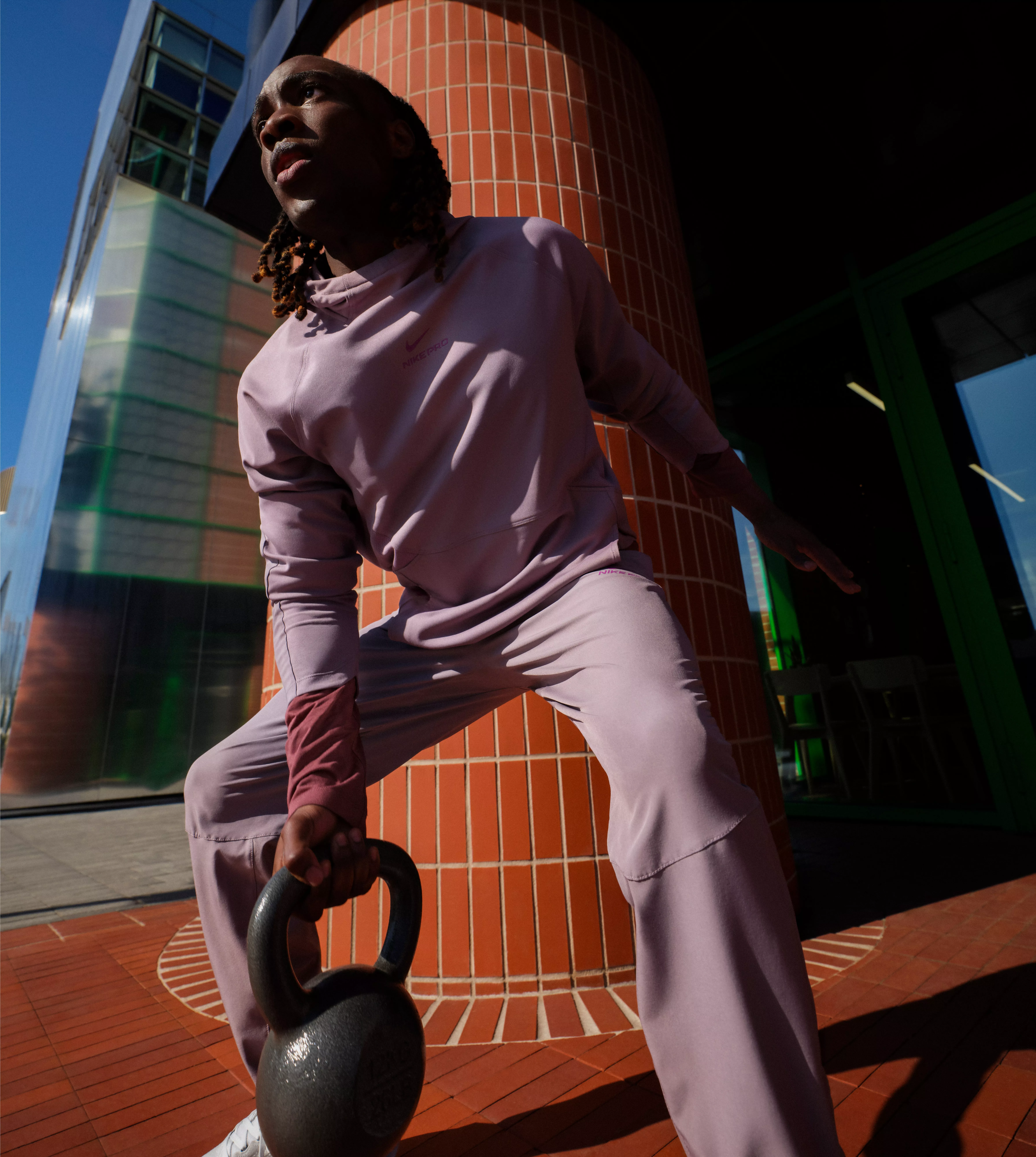 Athlete in mauve tracksuit lifting a kettlebell outdoors by a modern building, strength training in sunlight
