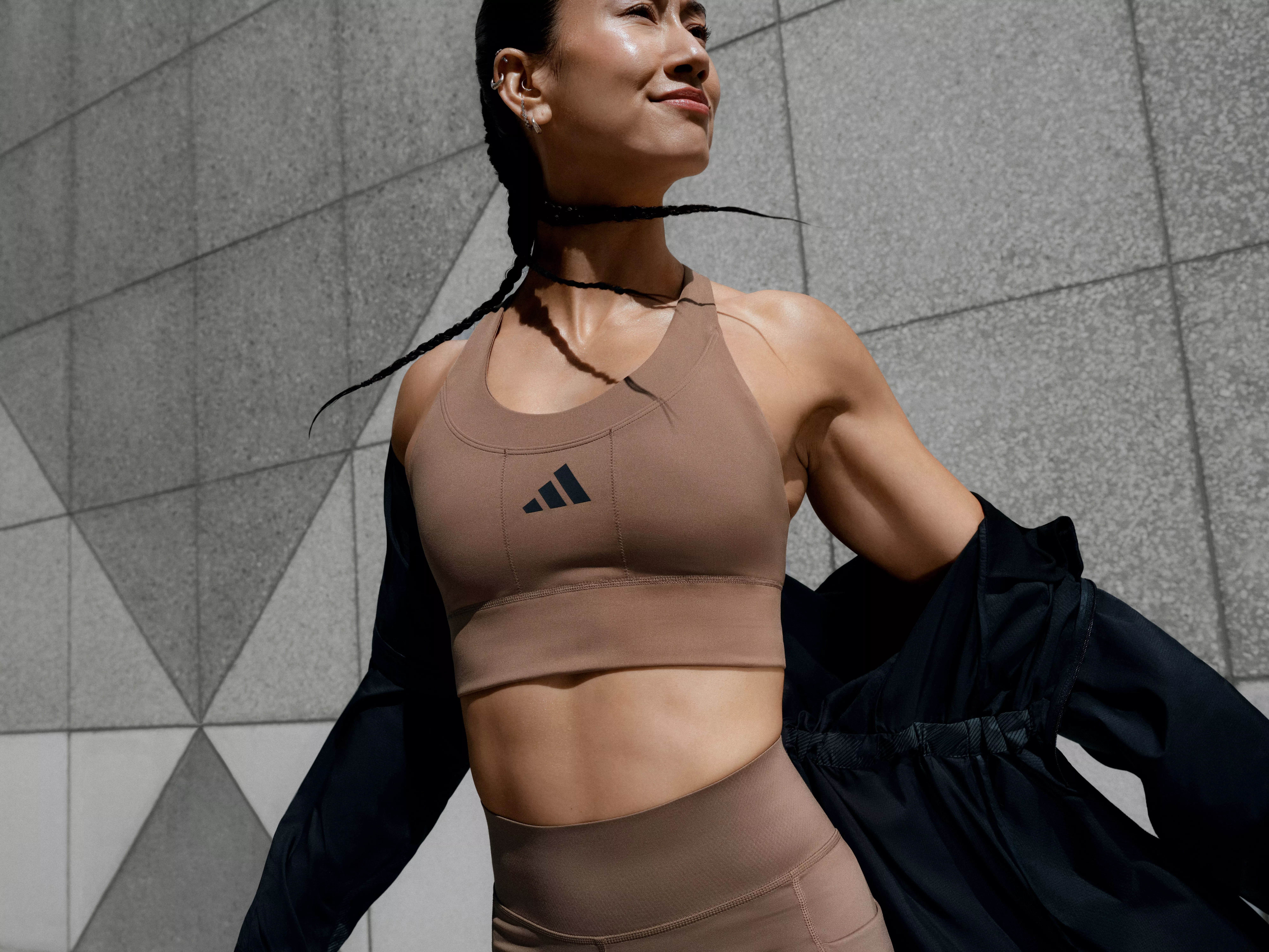 Athletic woman in brown sports bra and leggings, outdoors by concrete wall, confident fitness lifestyle look