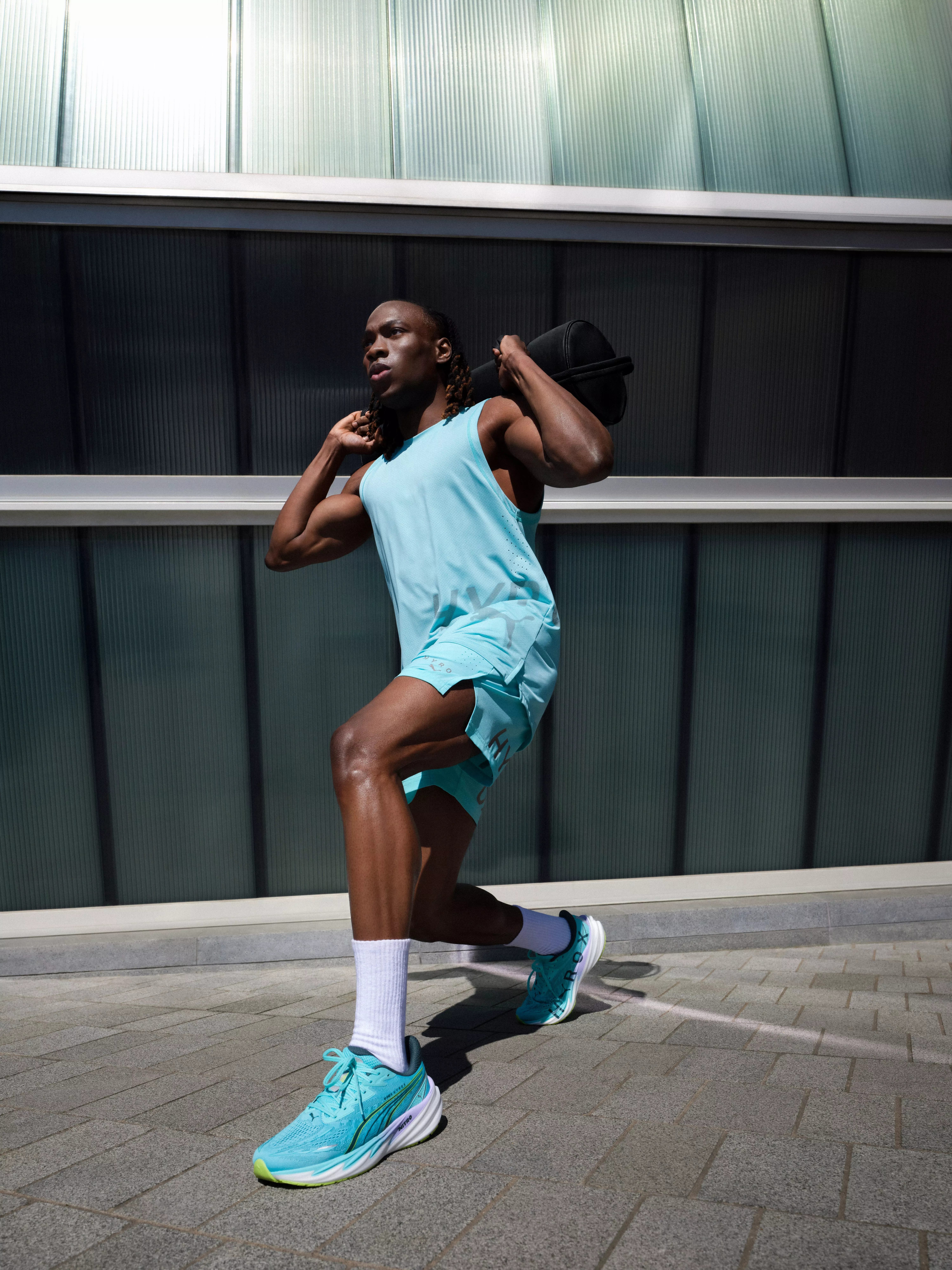 Athlete doing forward lunge with sandbag outdoors, wearing blue sportswear and running trainers in sunlight