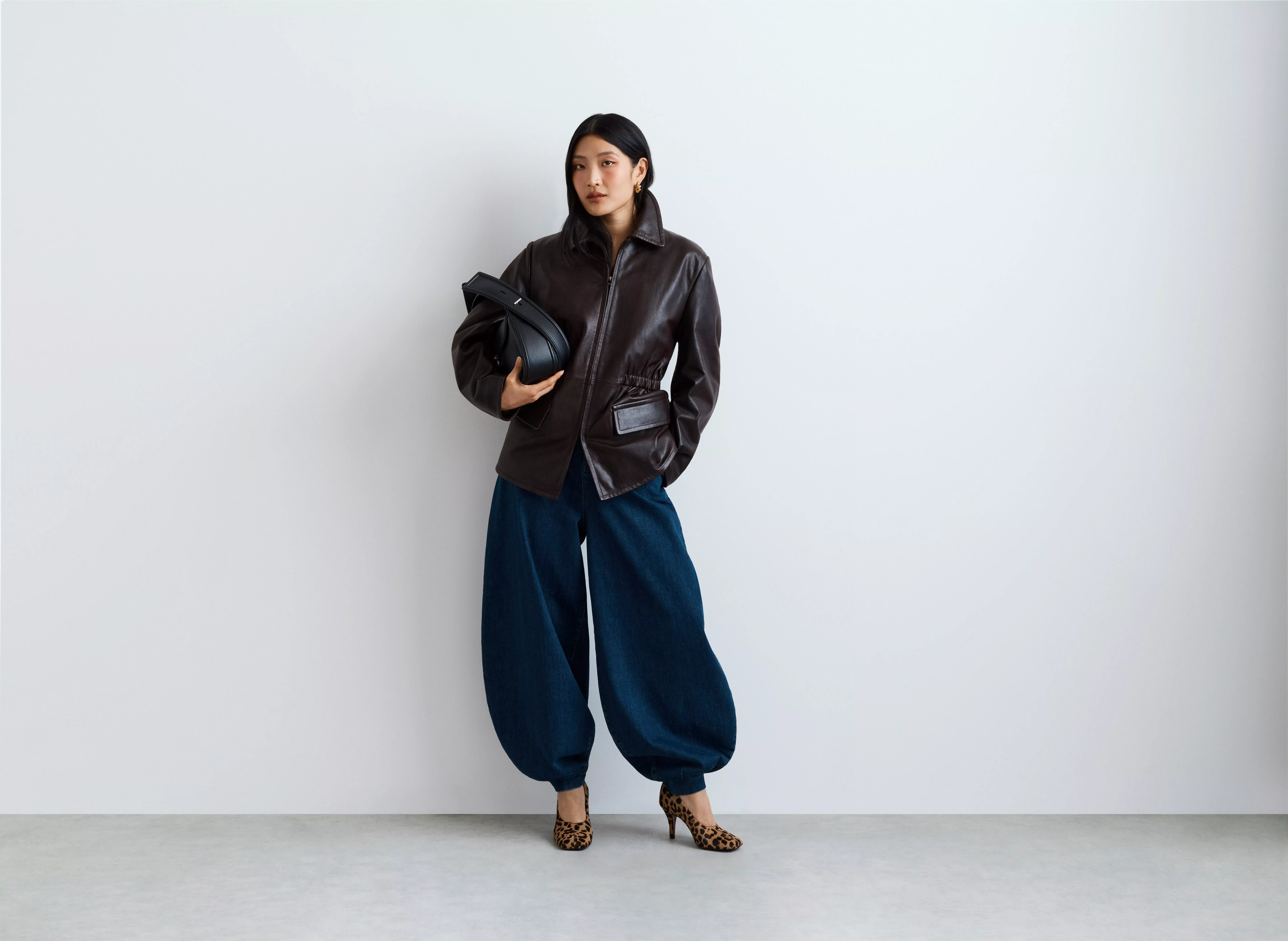 Fashion model in brown leather jacket, wide-leg denim trousers, holding black bag, wearing leopard heels in studio.
