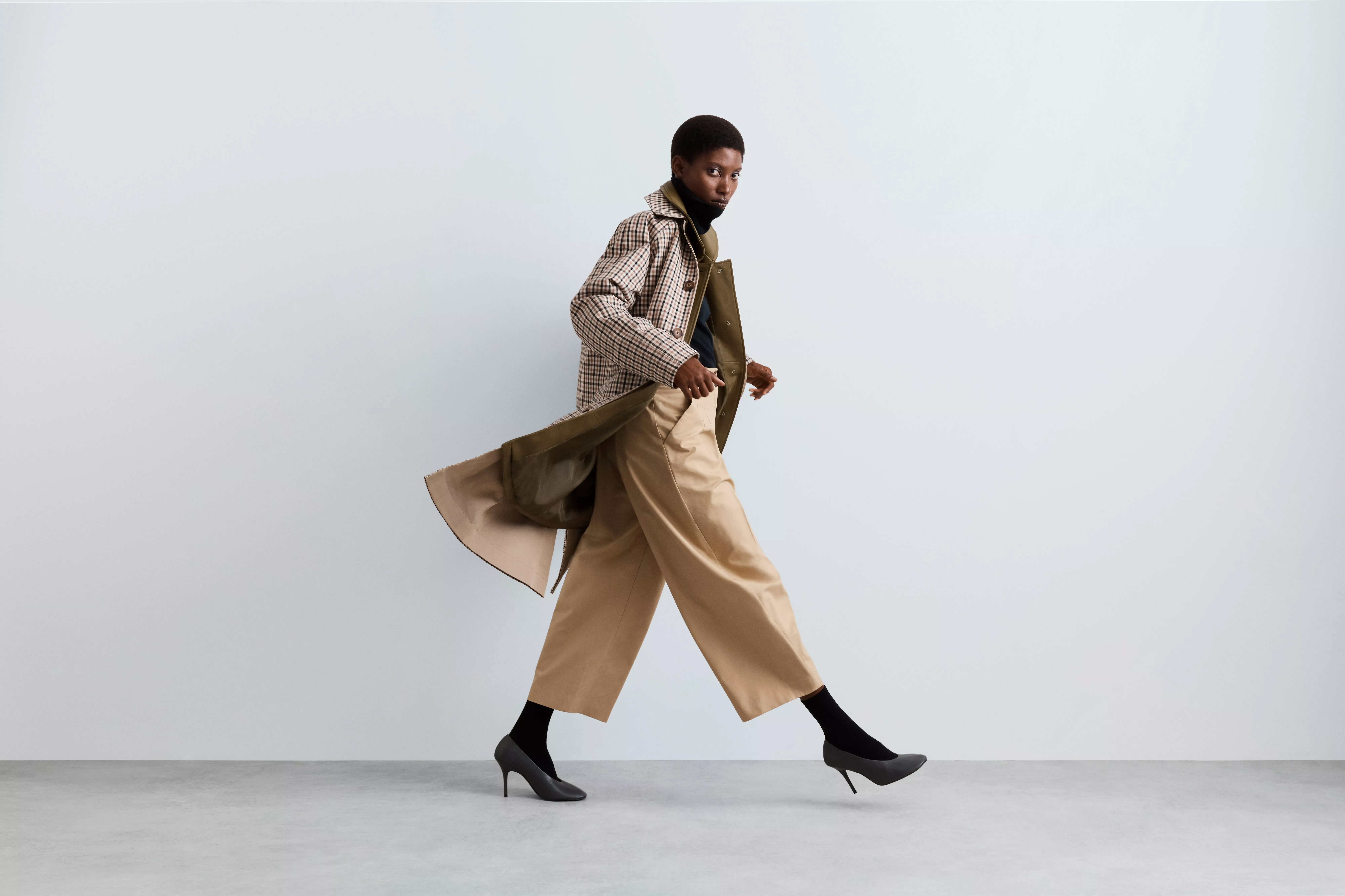 Fashion model walking in studio wearing checked coat, wide-leg beige trousers and black heeled boots