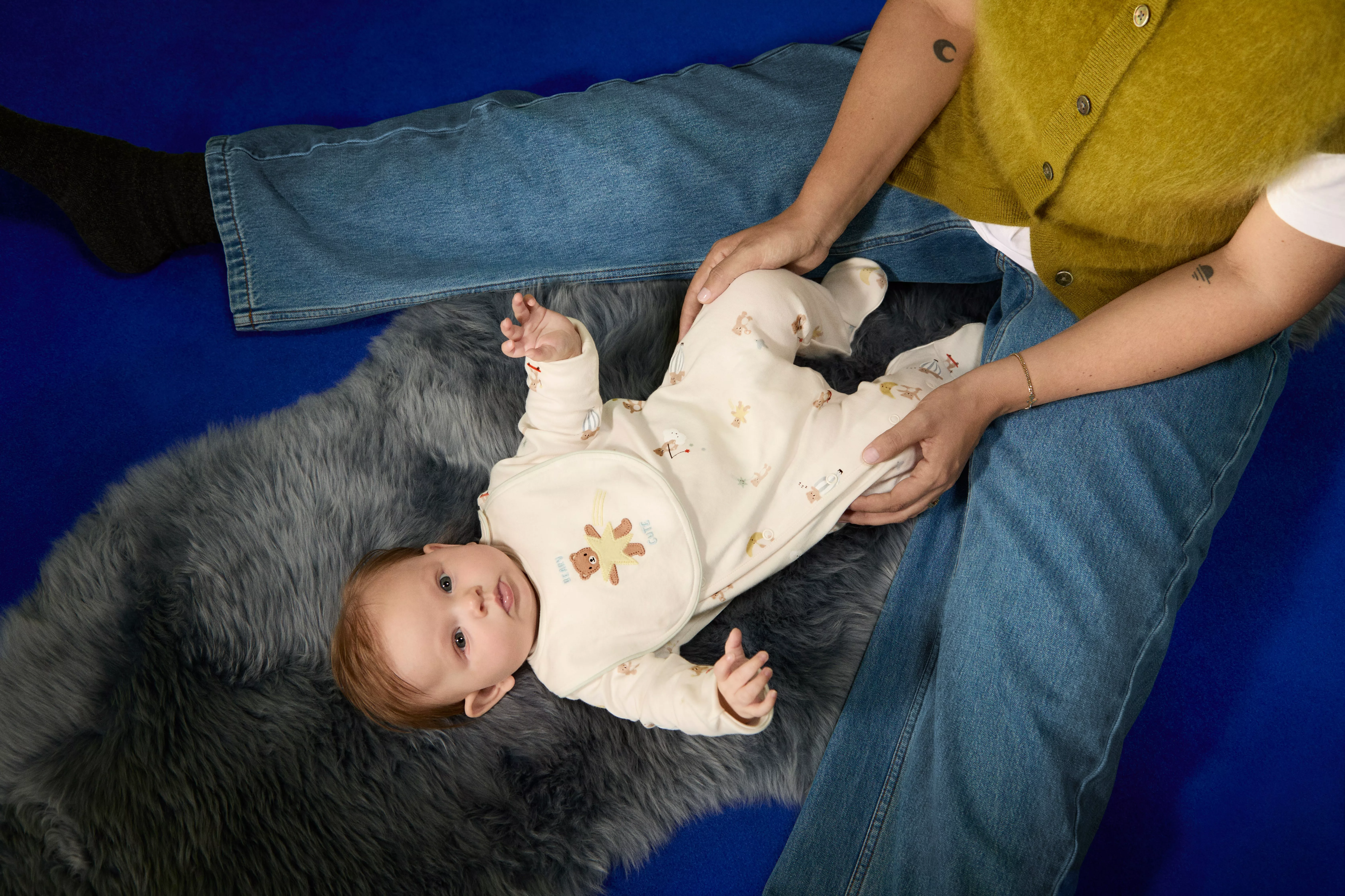 Baby lying on grey faux fur rug with parent holding legs, wearing cream sleepsuit and bib on blue floor