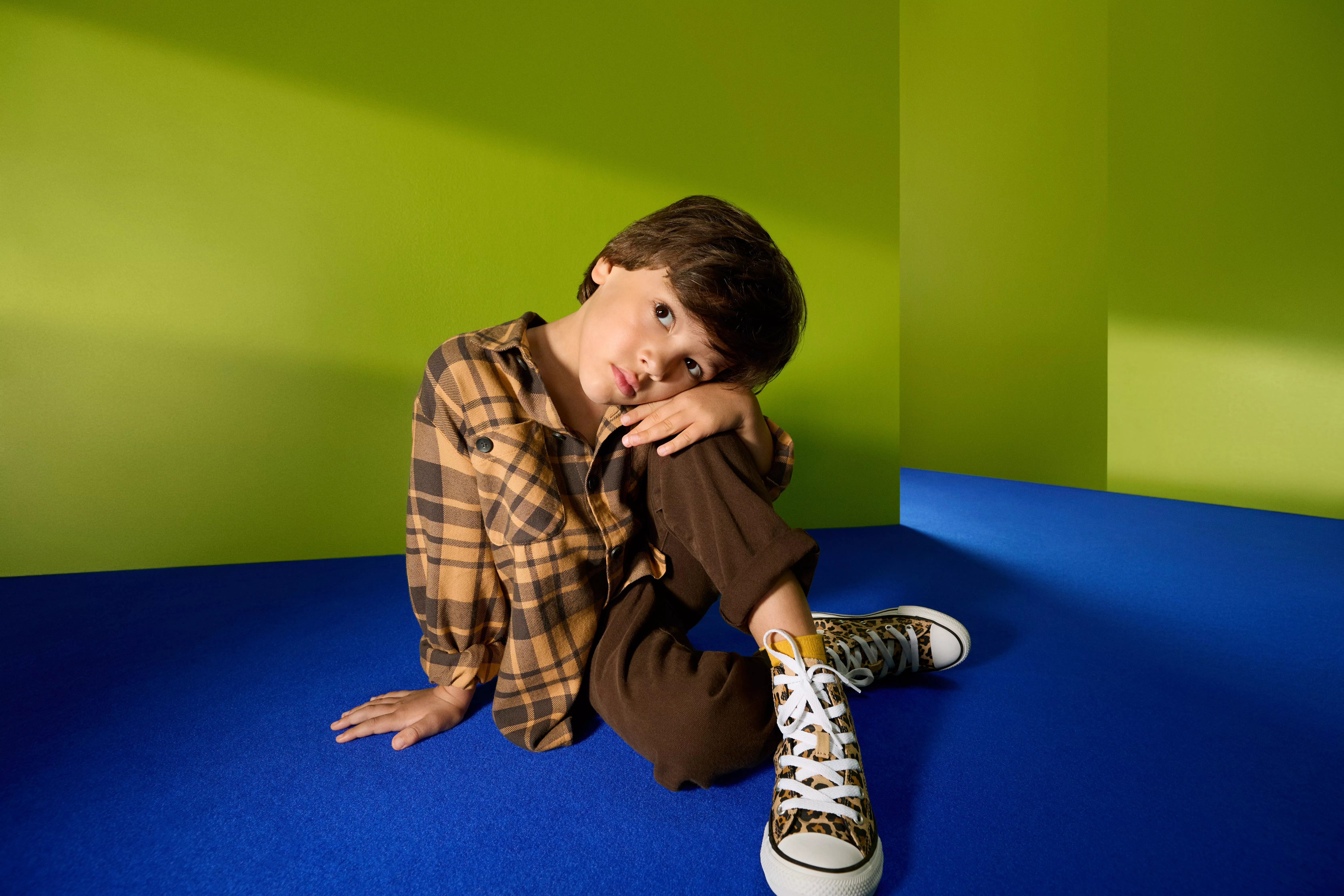 Young boy in checked shirt and brown trousers sitting on blue floor, wearing leopard print trainers, green backdrop