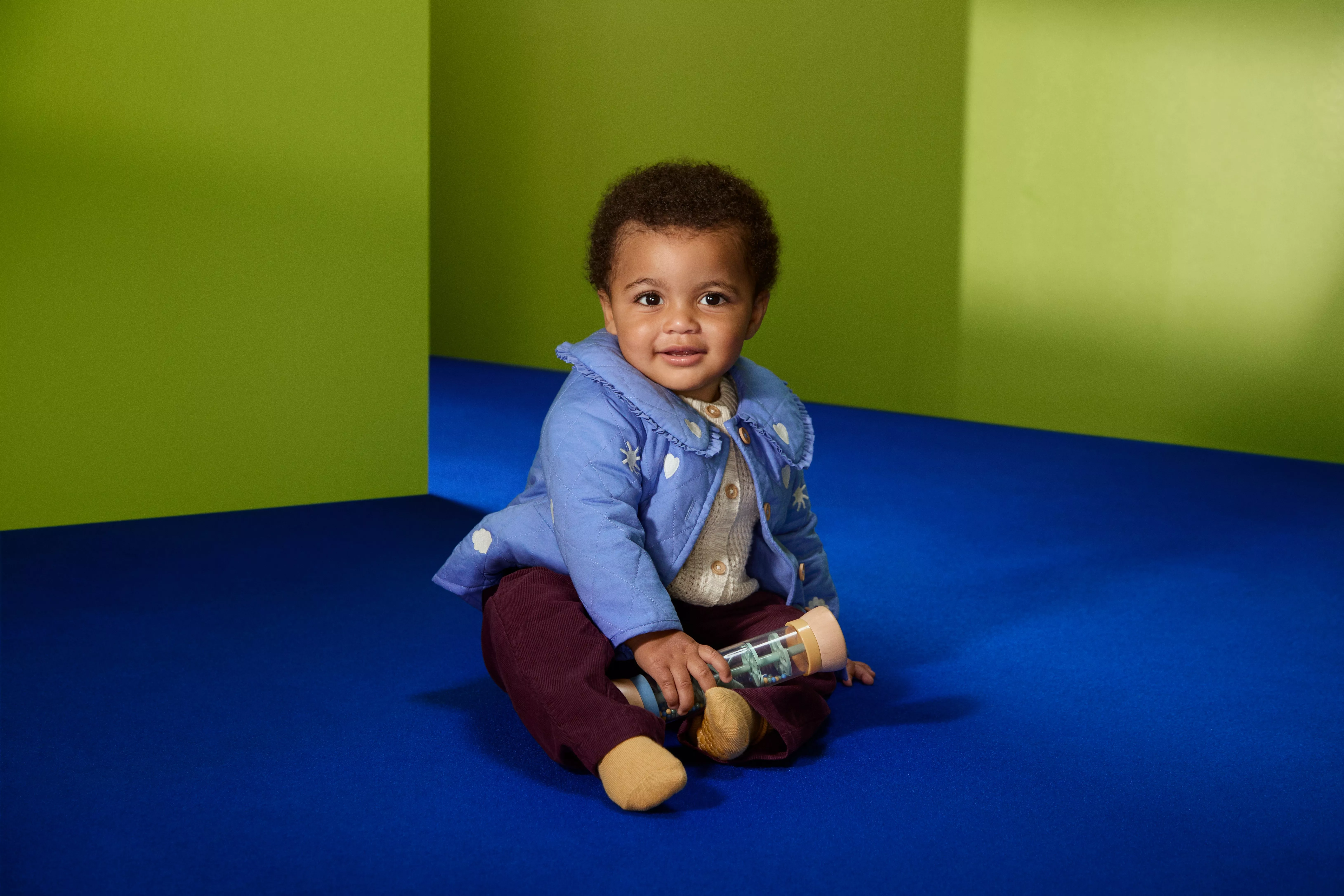 Smiling baby in blue jacket sitting on blue floor, holding toy bottle against green studio backdrop