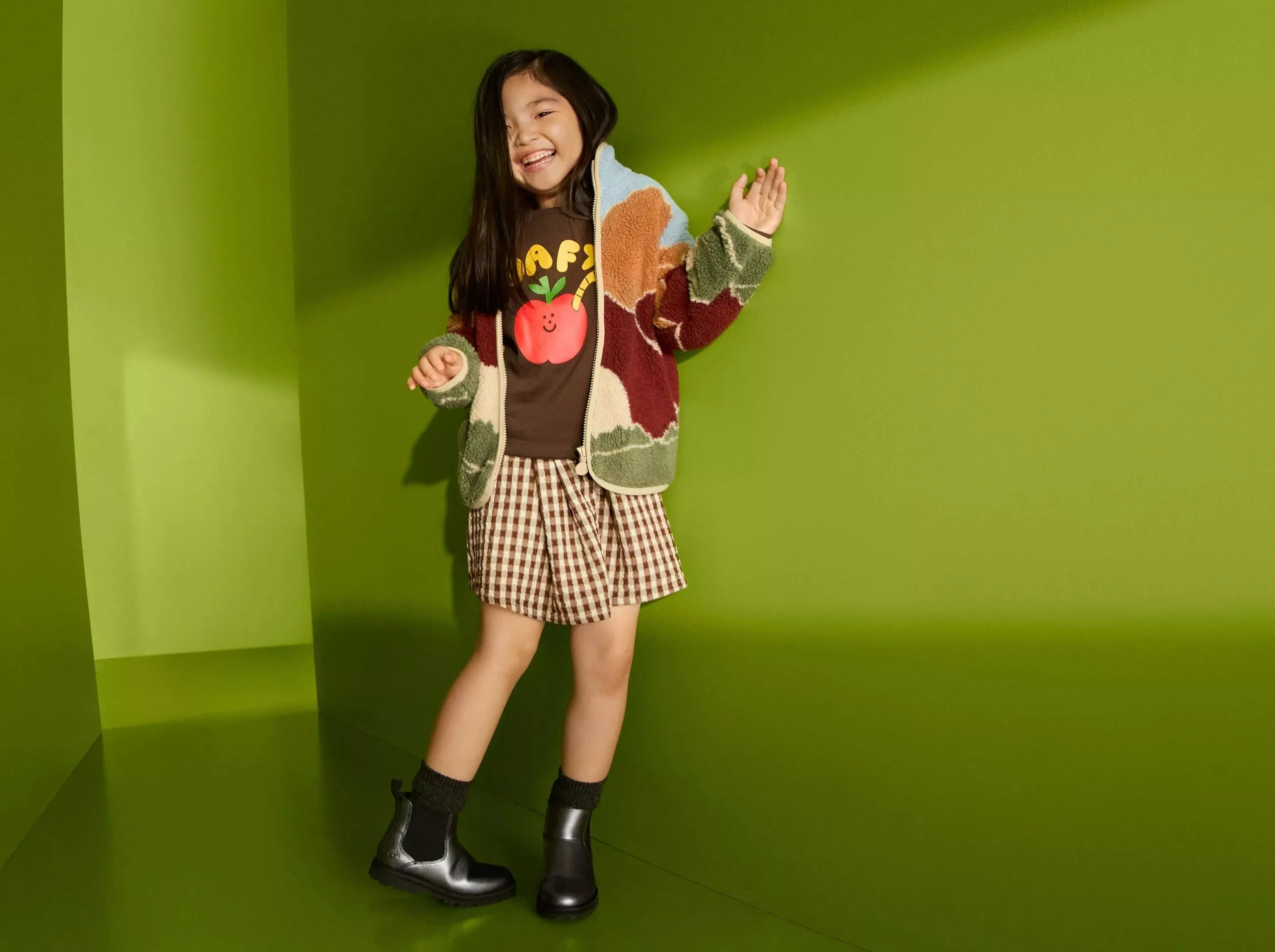 Smiling girl in cosy fleece jacket, apple graphic top and gingham shorts, posing against green studio wall