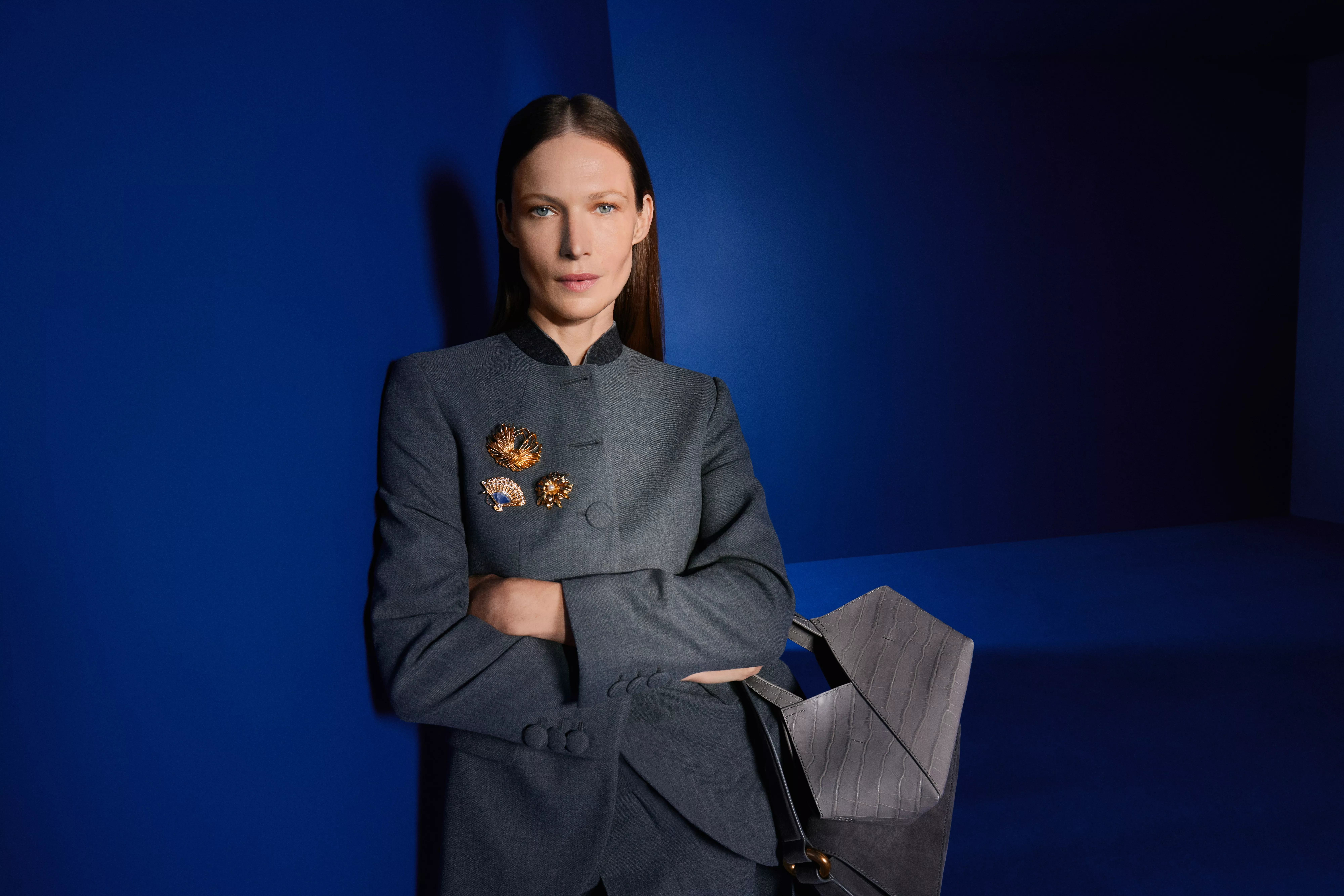 Fashion portrait of woman in grey blazer with gold brooches, holding designer leather bag against blue backdrop