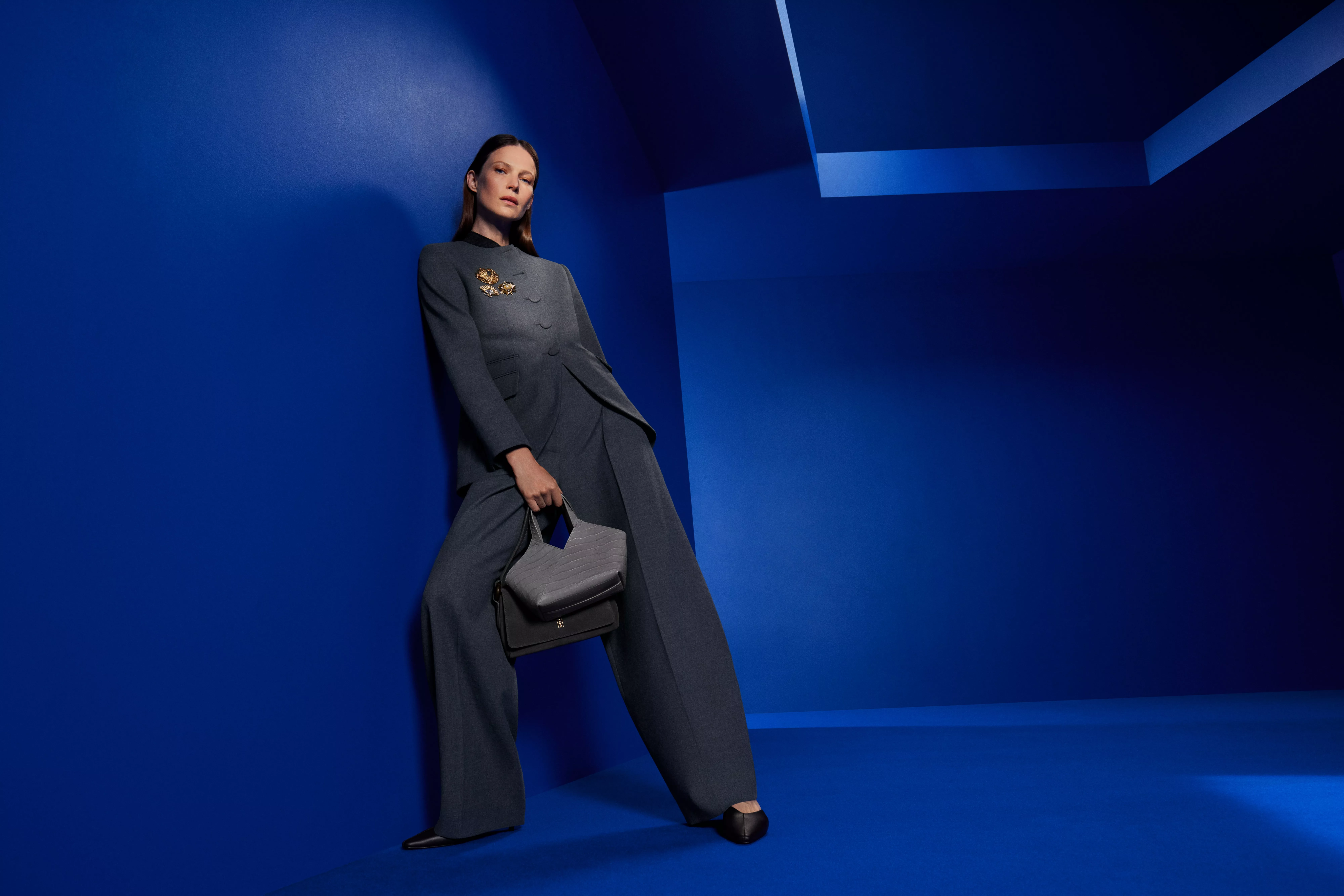 Fashion model in grey tailored suit holding designer handbag in modern blue studio, editorial style photo