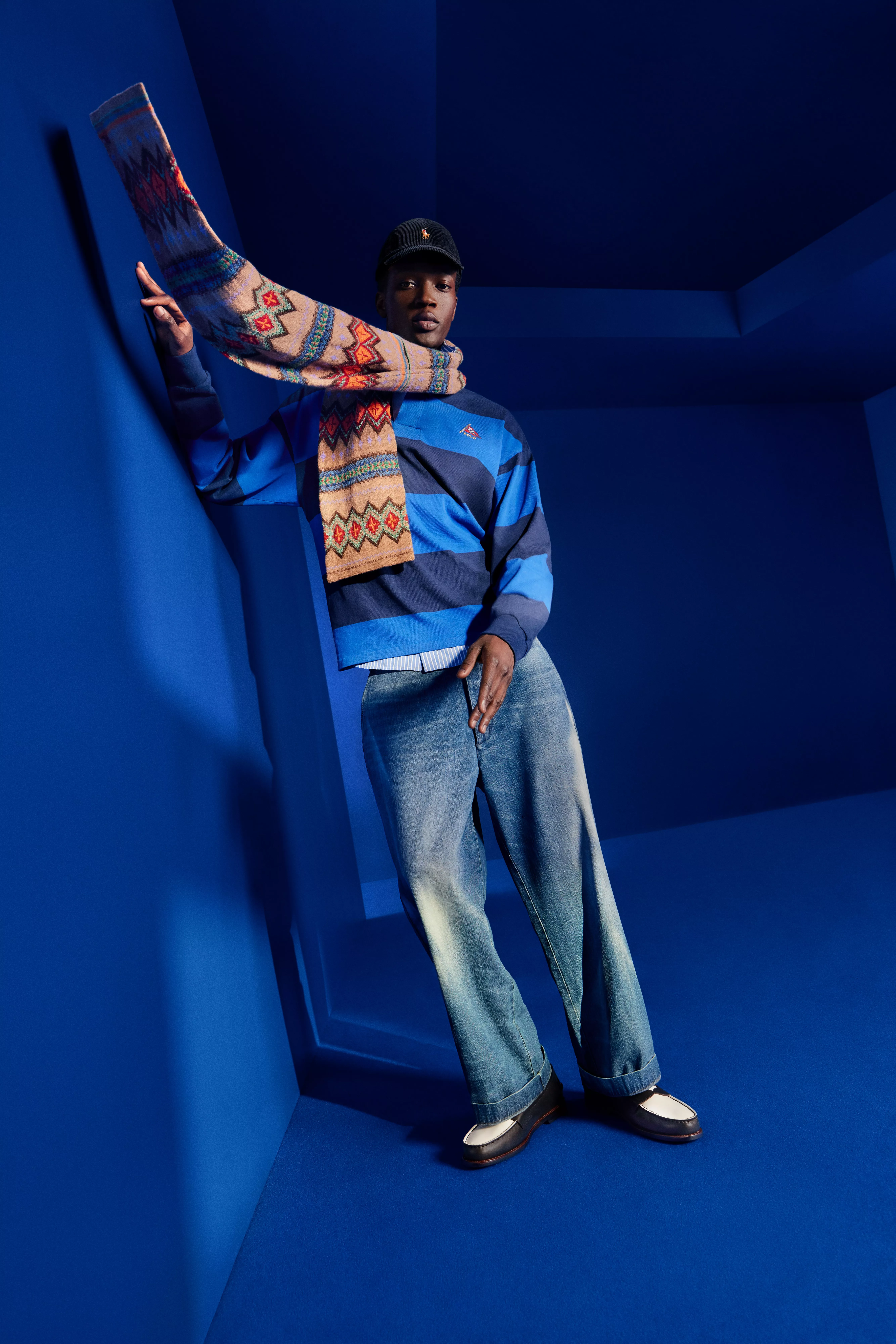 Fashion portrait of man in blue striped rugby top, jeans and patterned scarf, posing in blue studio set