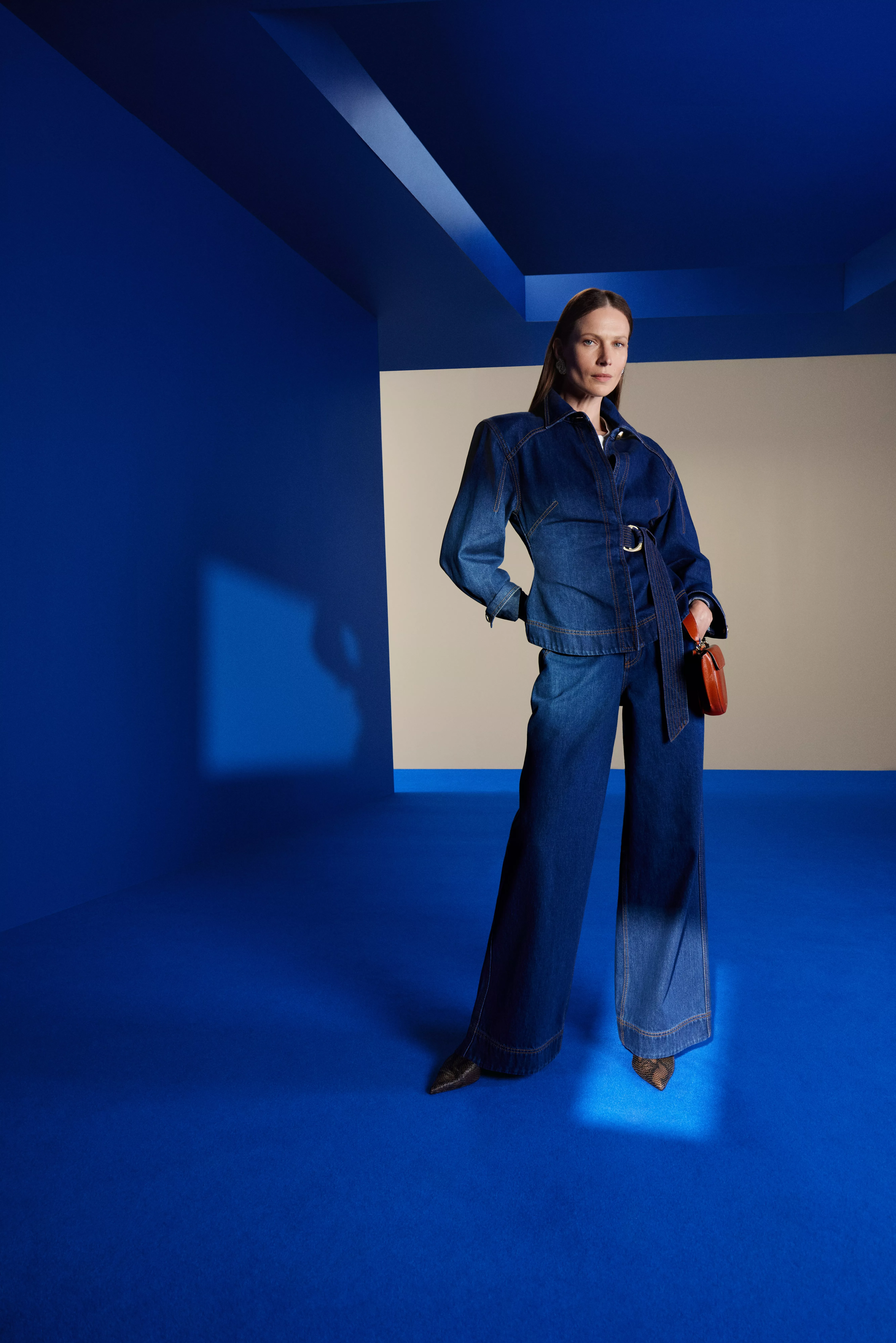 Fashion model in belted denim jacket and wide-leg jeans, standing in bold blue studio with red handbag