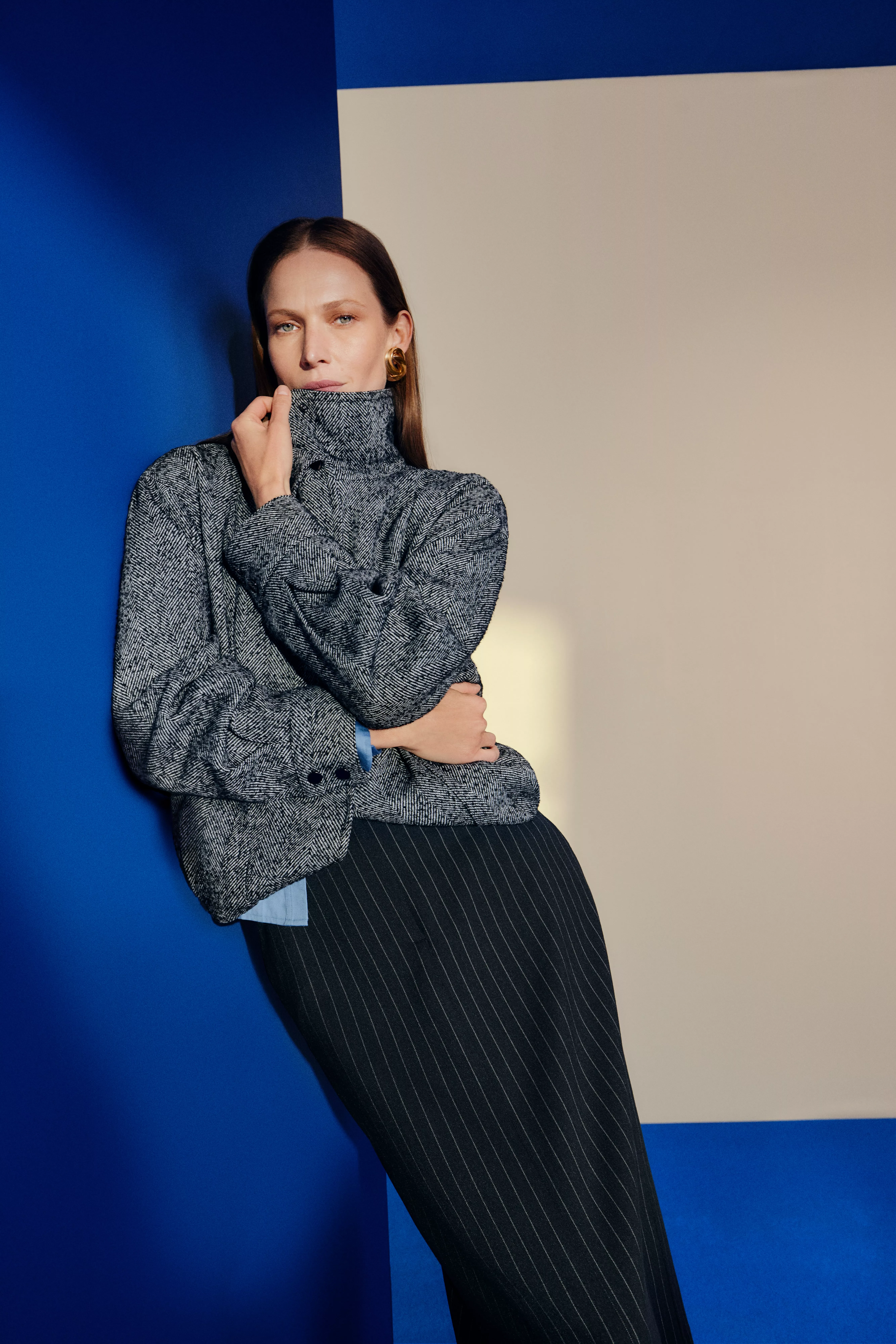 Fashion portrait of woman in grey herringbone jacket and pinstripe trousers against blue and beige wall
