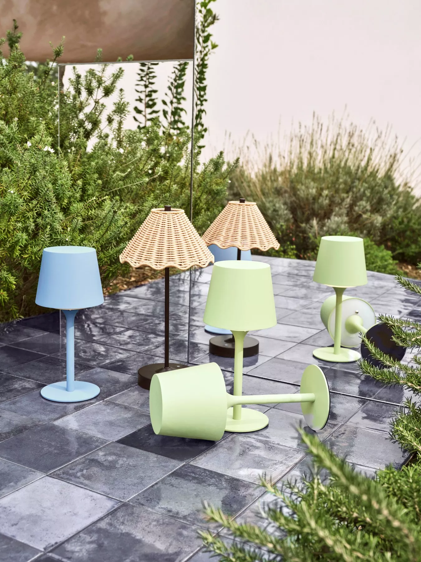 Colourful table lamps, including wicker shades, arranged on a tiled outdoor patio, surrounded by greenery.