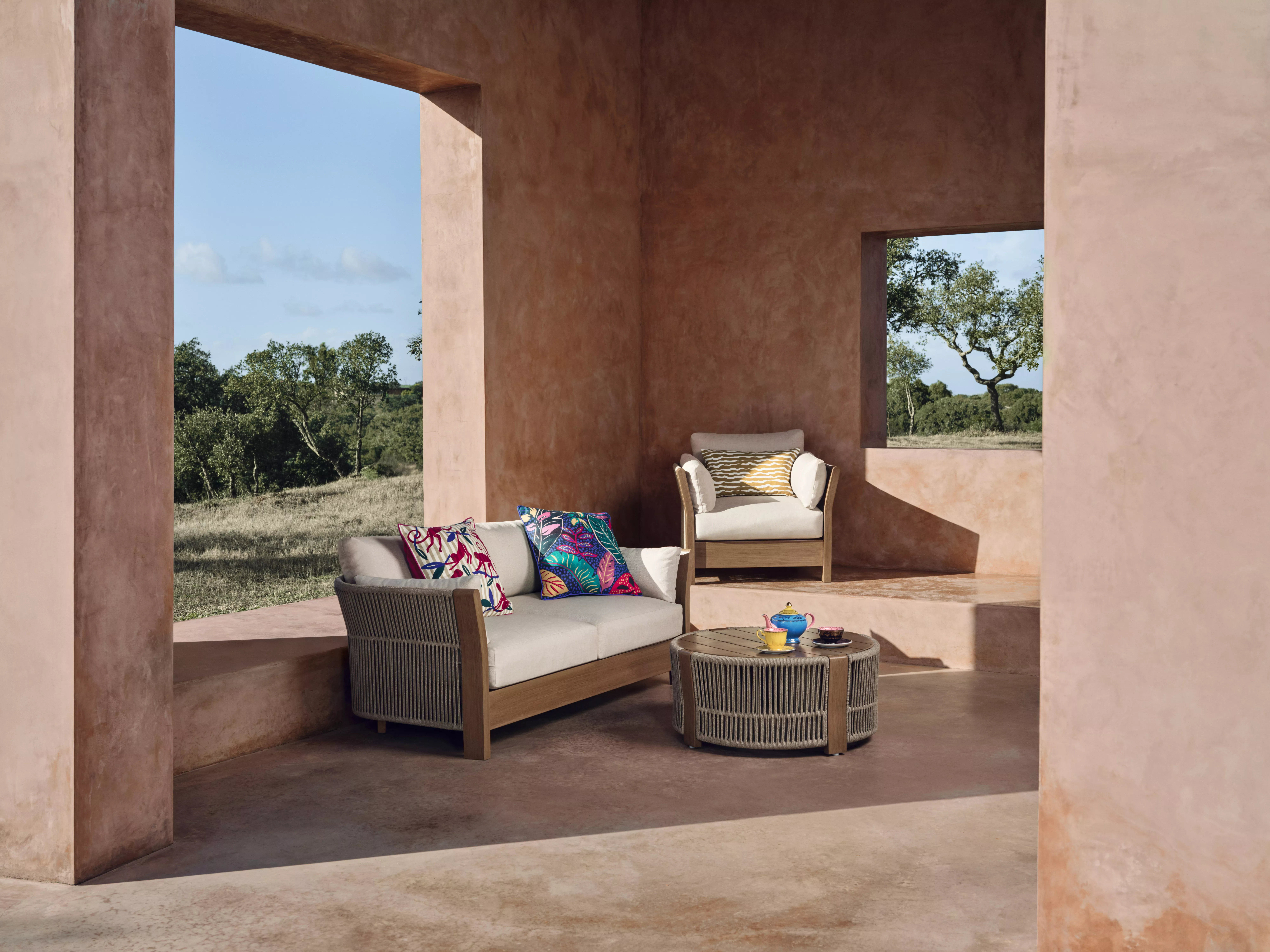 Two chairs and a table on a sandy beach overlook a vast, wavy ocean, surrounded by coastal plants.