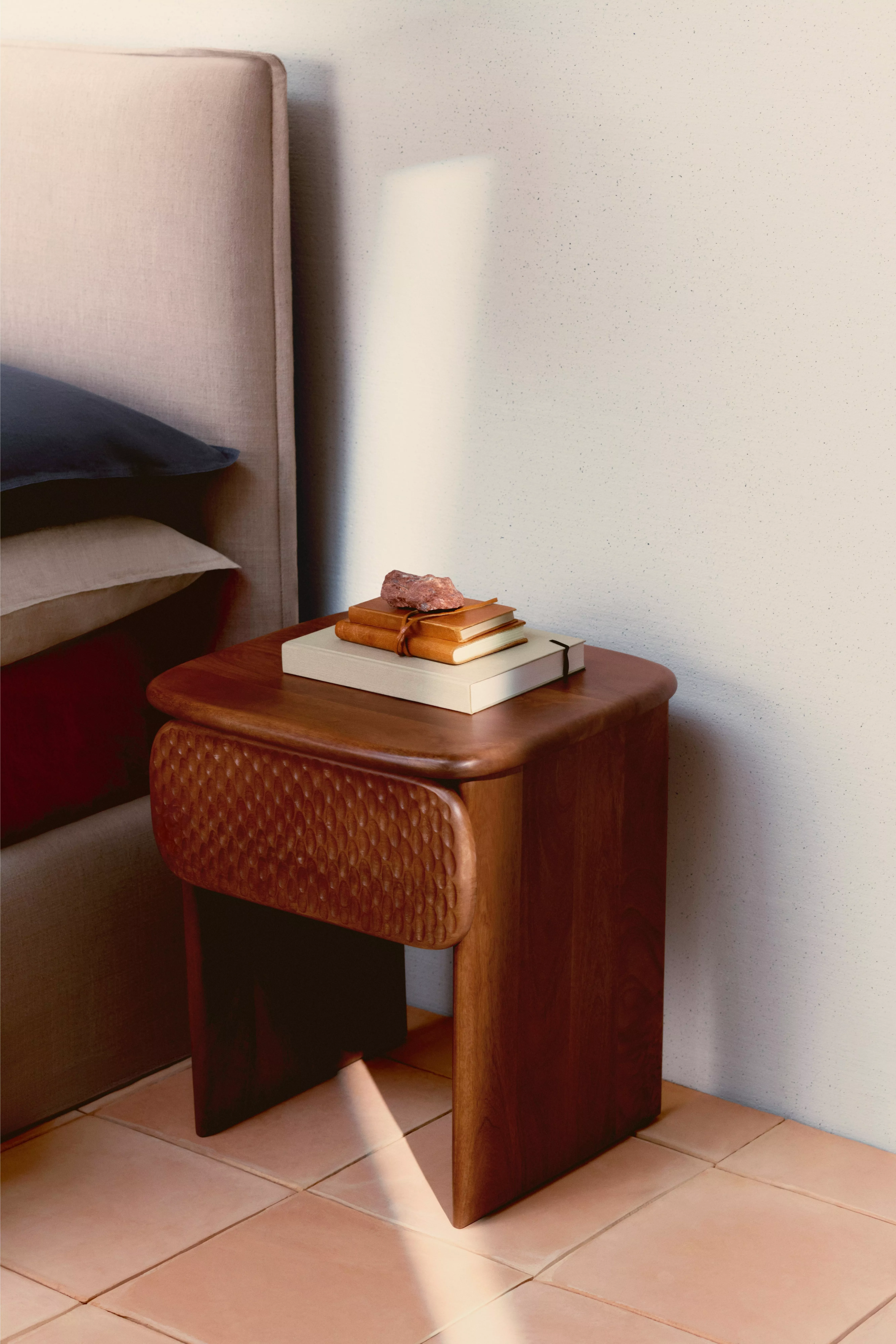 Mid-century wooden bedside table beside upholstered bed, with stacked books and soft morning light on tiled floor