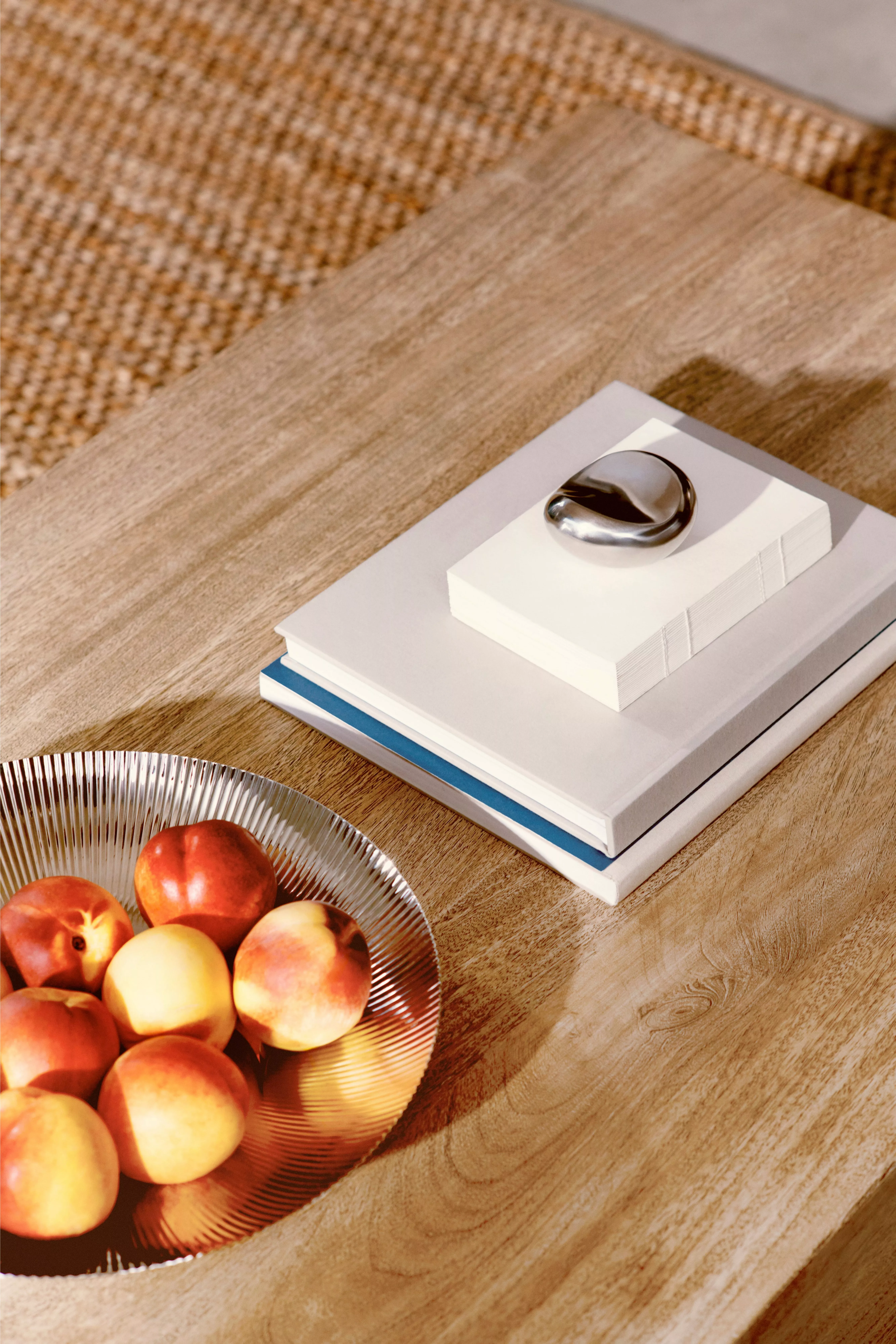 Sunlit wooden coffee table with stacked books, chrome paperweight and glass bowl of peaches in modern living room