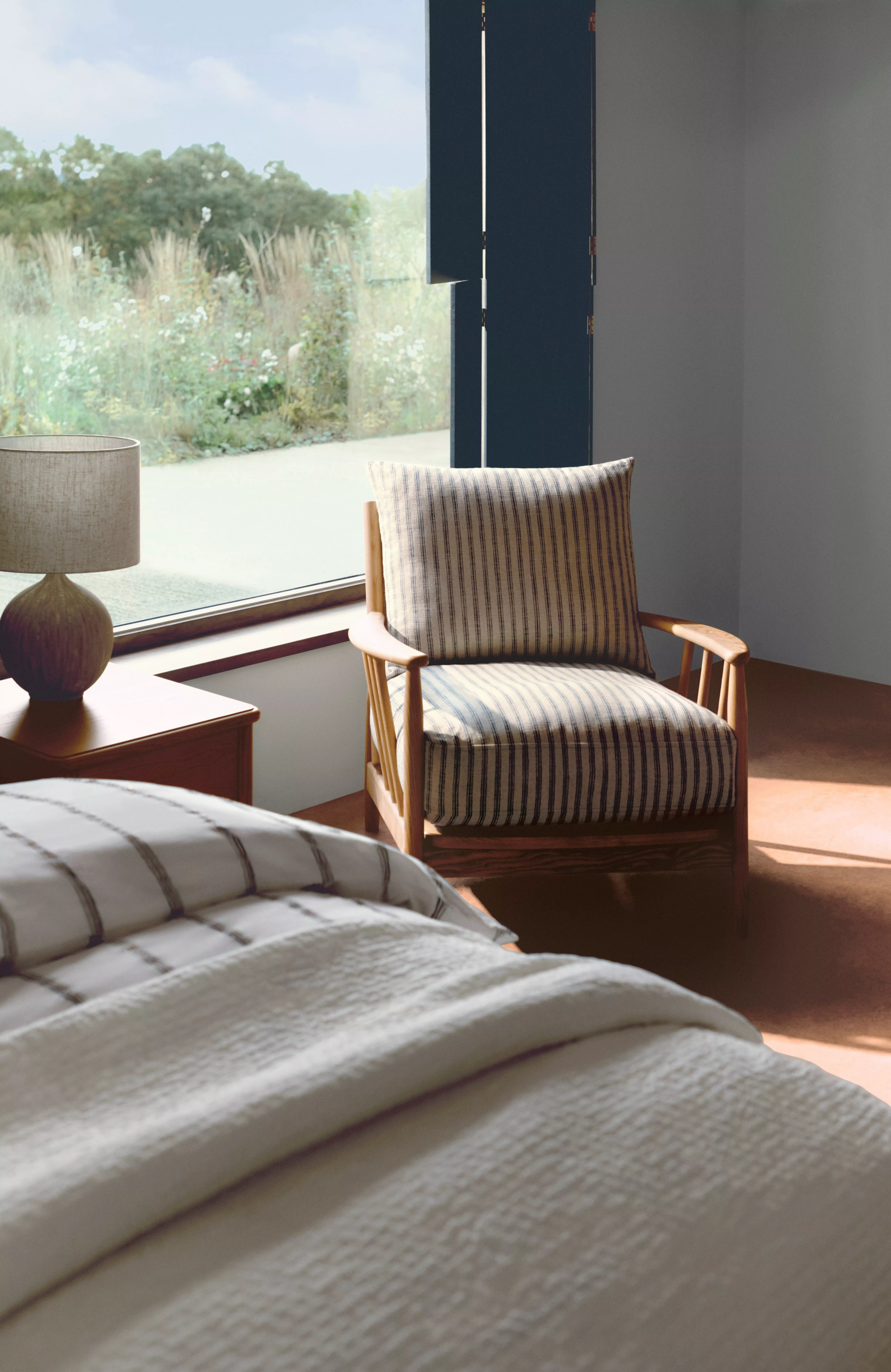 Sunlit modern bedroom with striped armchair, bedside table and lamp beside large window overlooking garden.