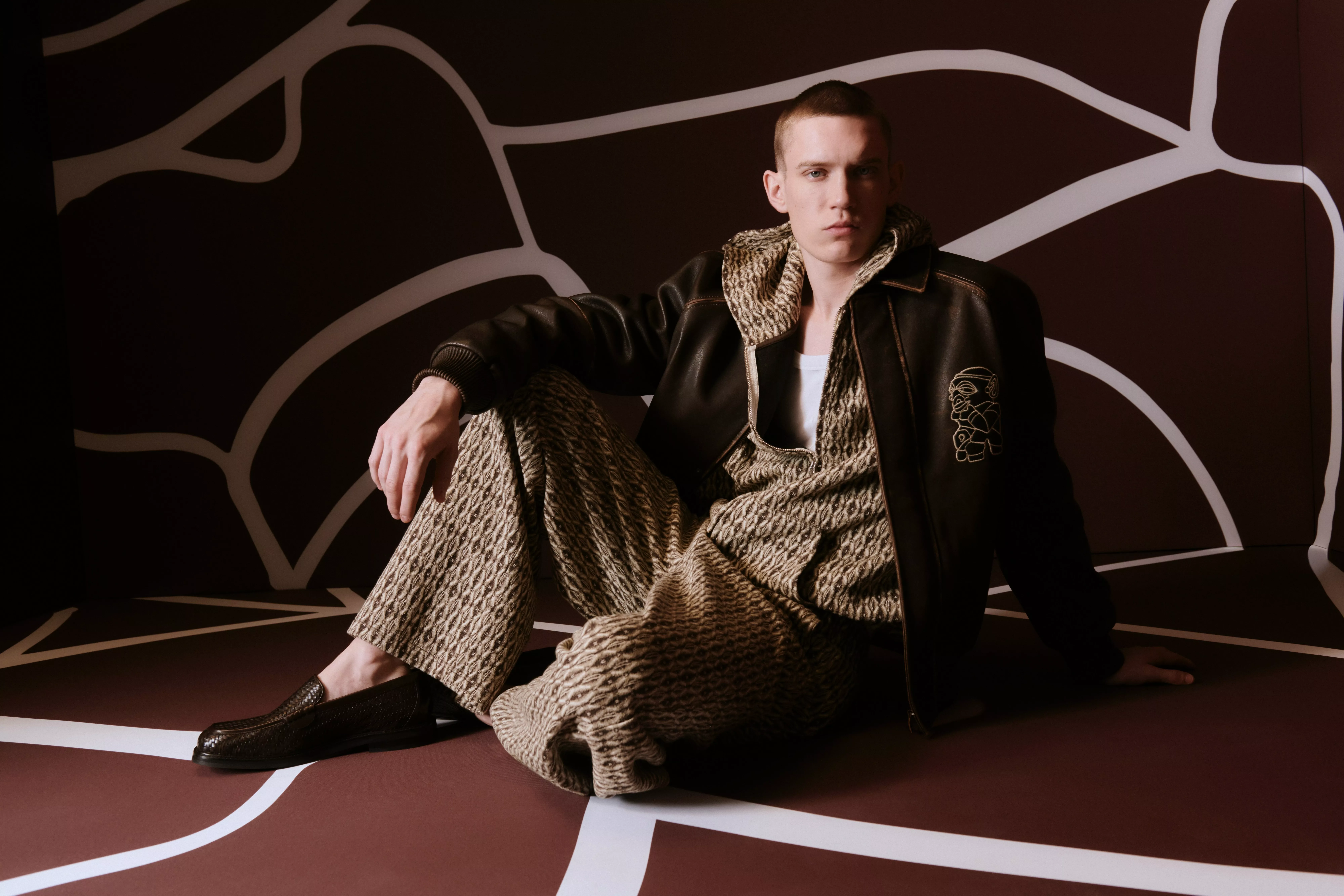 Model in a textured outfit and black jacket sits against a geometric backdrop, exuding a stylish and relaxed vibe.