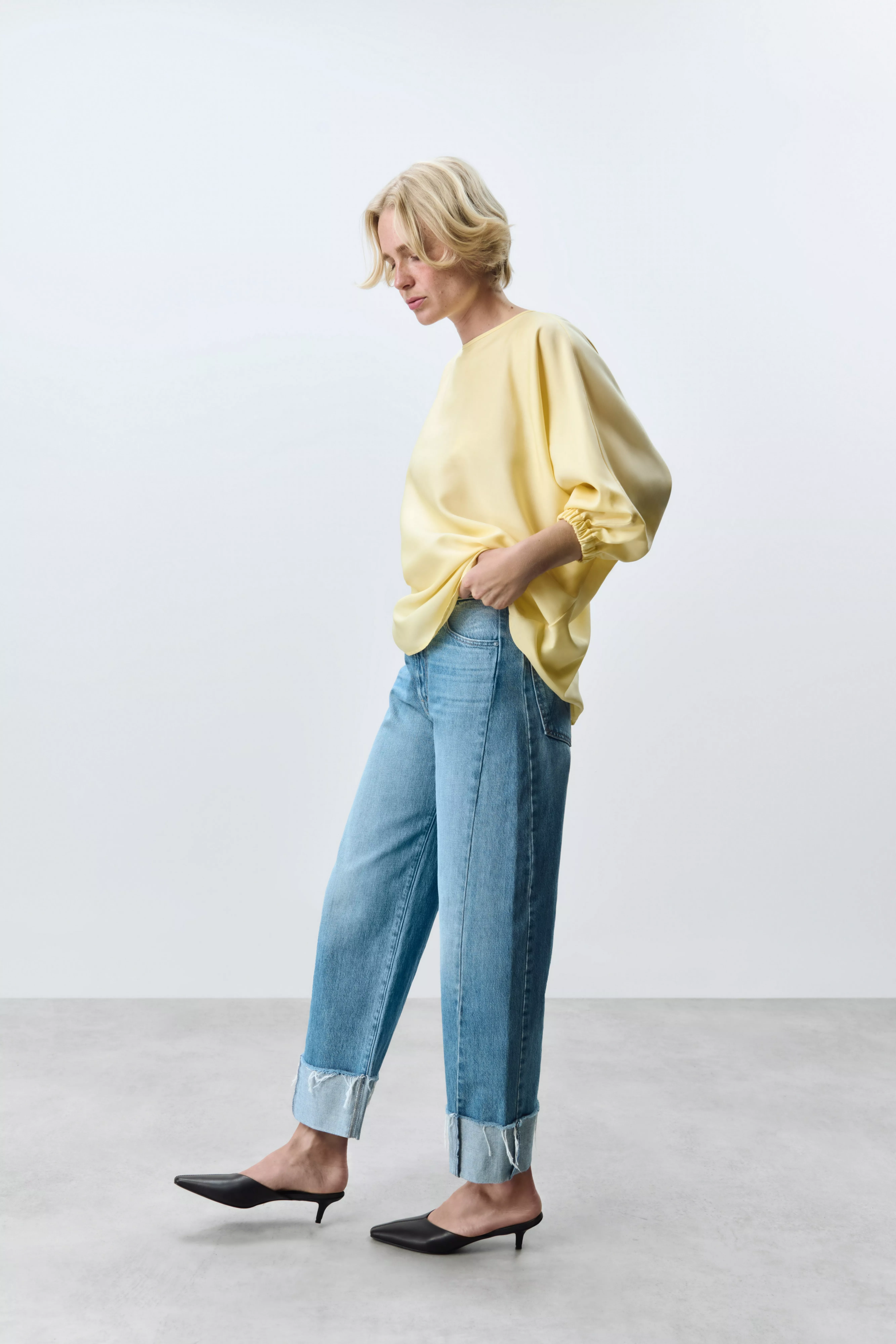 Woman in yellow satin blouse and cuffed blue jeans with black kitten heels, minimalist studio fashion shot