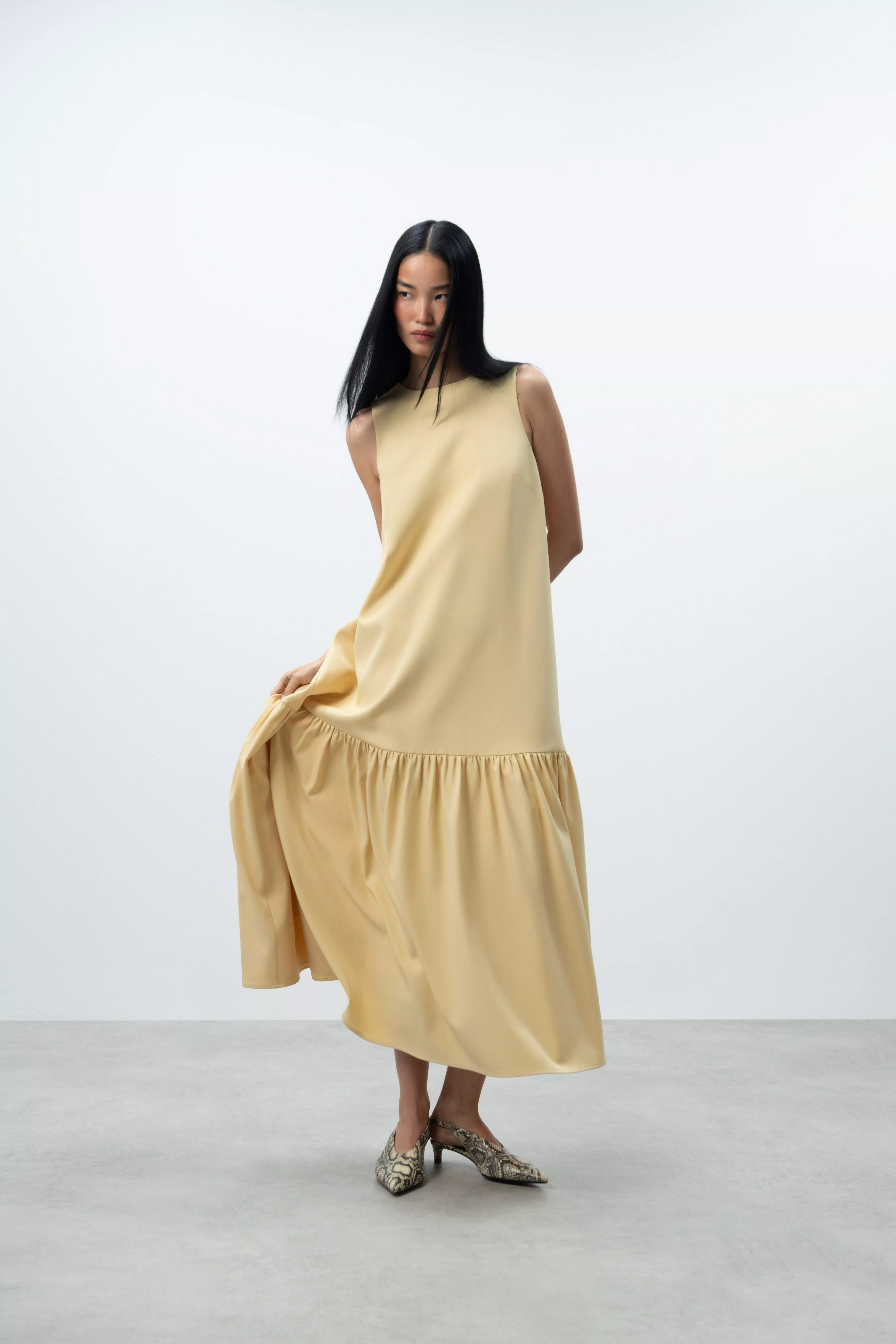 Model wearing a pale yellow sleeveless midi dress with gathered hem and snakeskin heels in a minimal studio setting