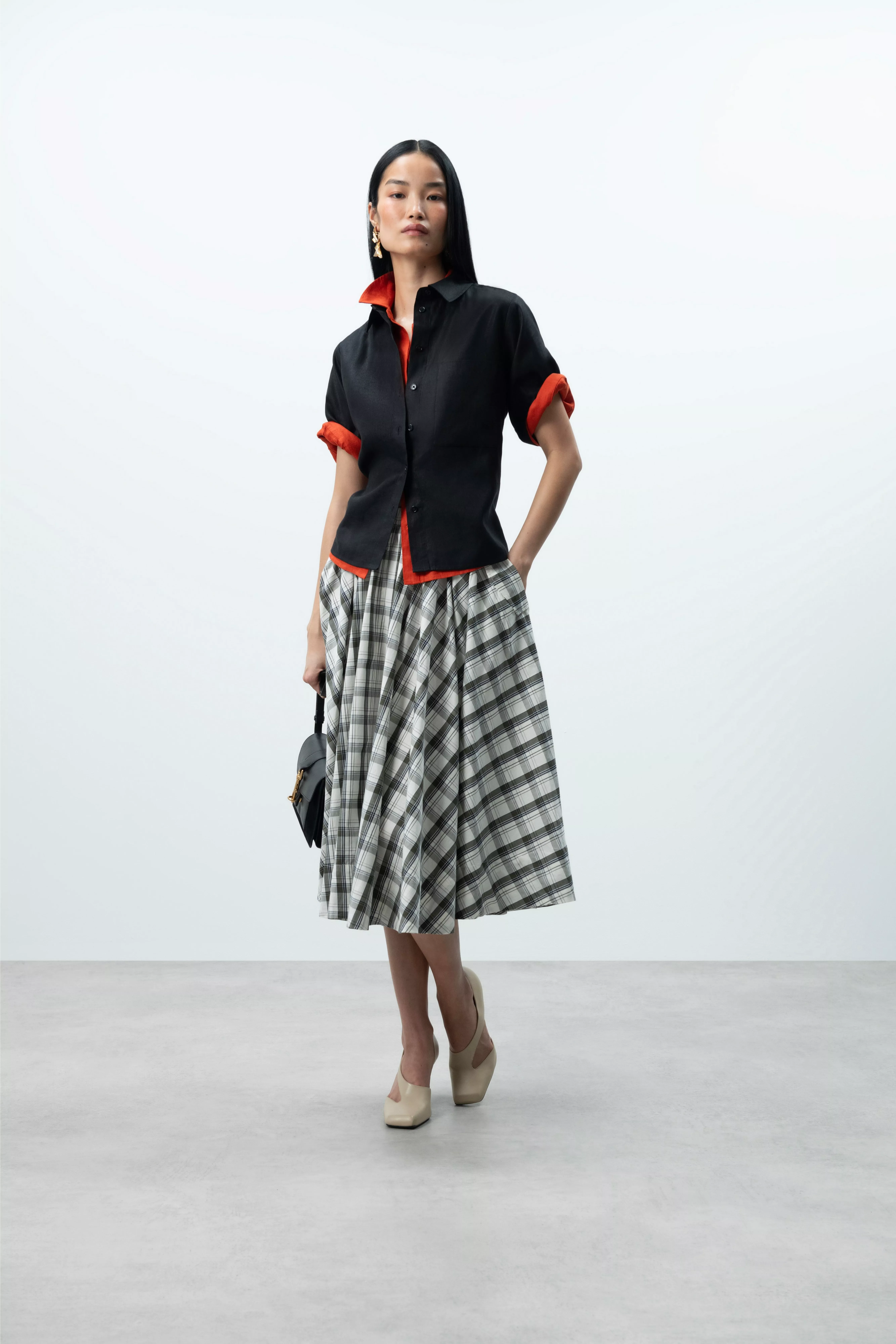 Studio fashion look: model in black shirt with red cuffs, grey plaid midi skirt, beige heels and handbag