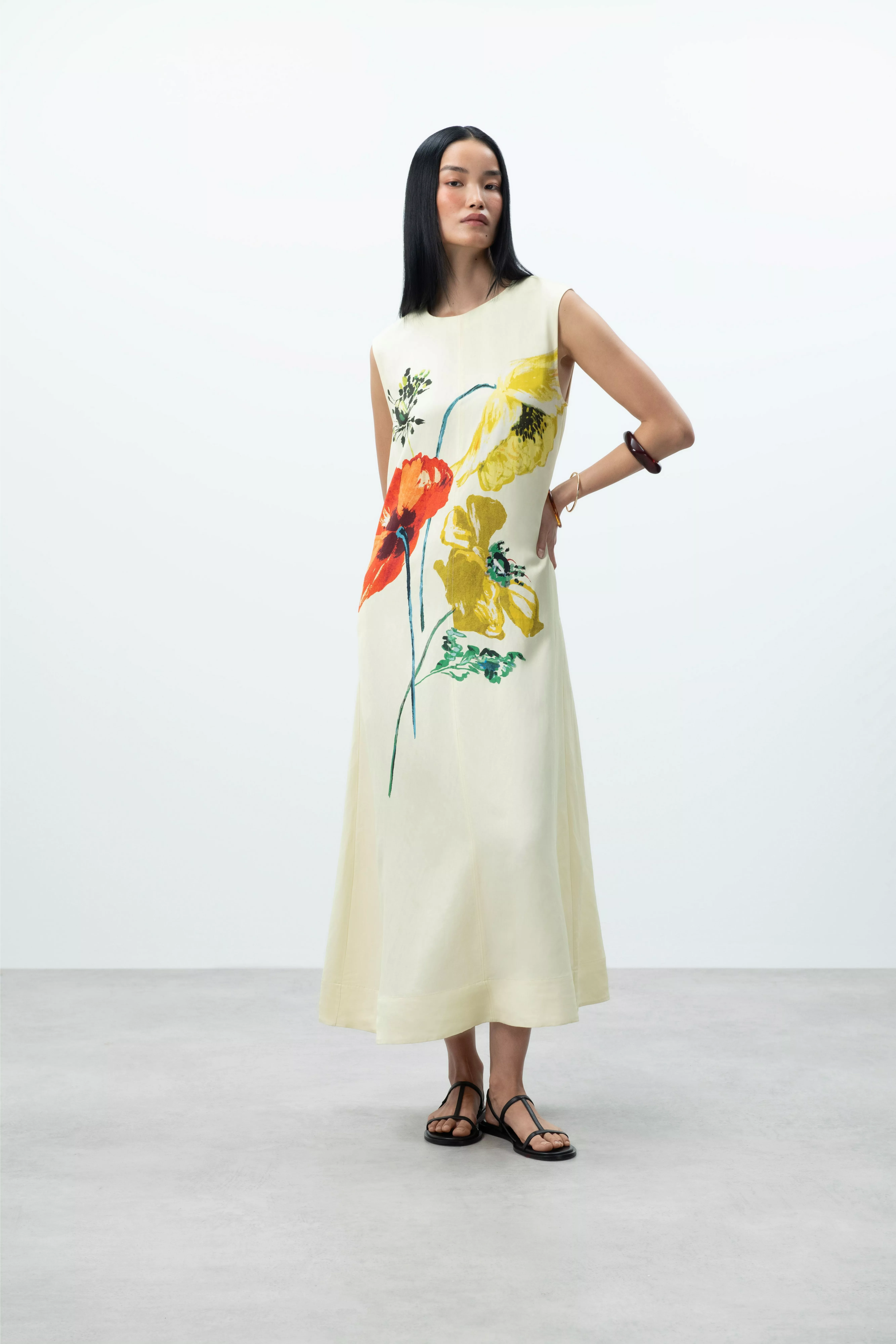 Model wearing a sleeveless cream midi dress with bold floral print, styled with black sandals in studio shot