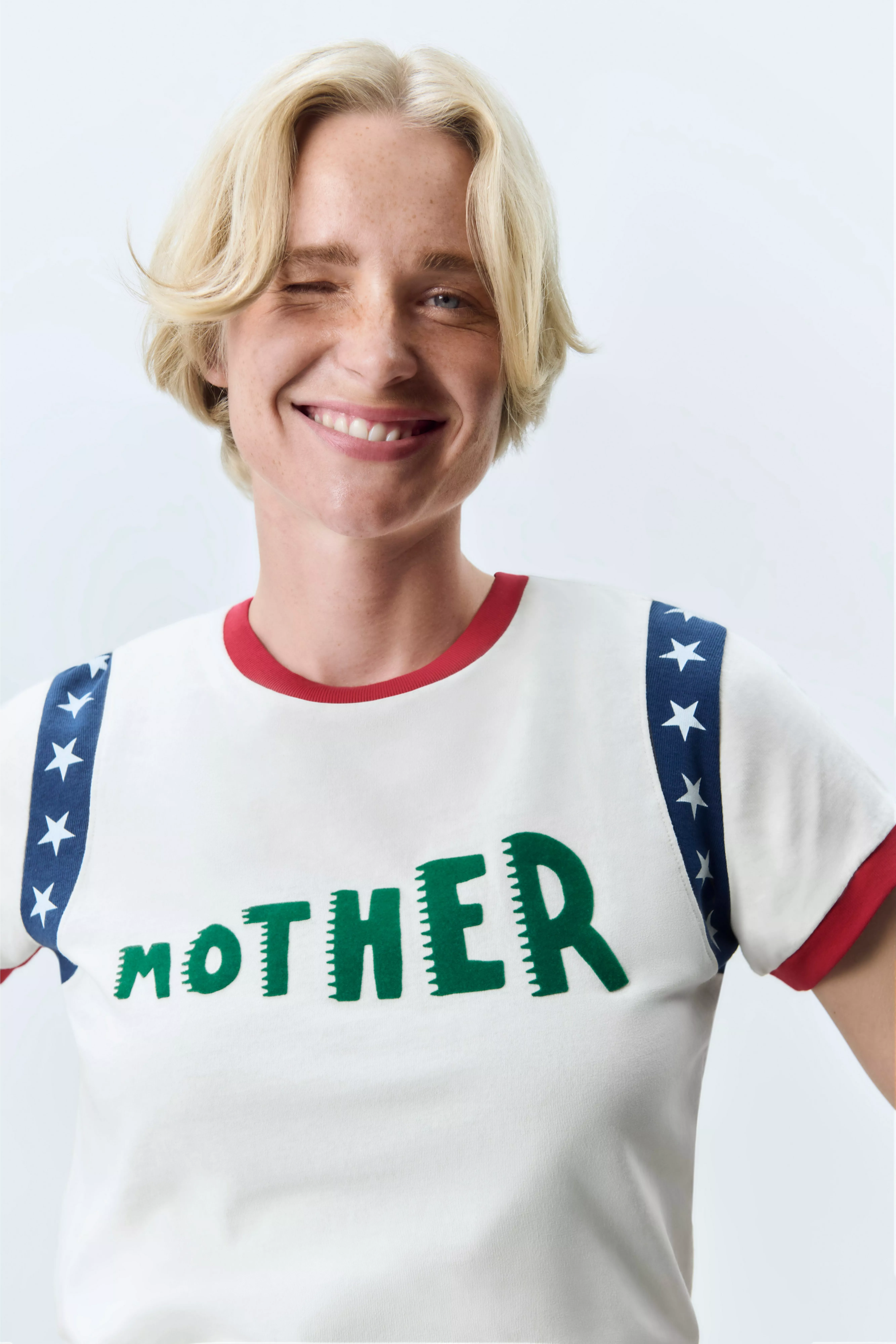 Smiling blonde person winking in “Mother” slogan T-shirt with star braces, studio portrait on white backdrop