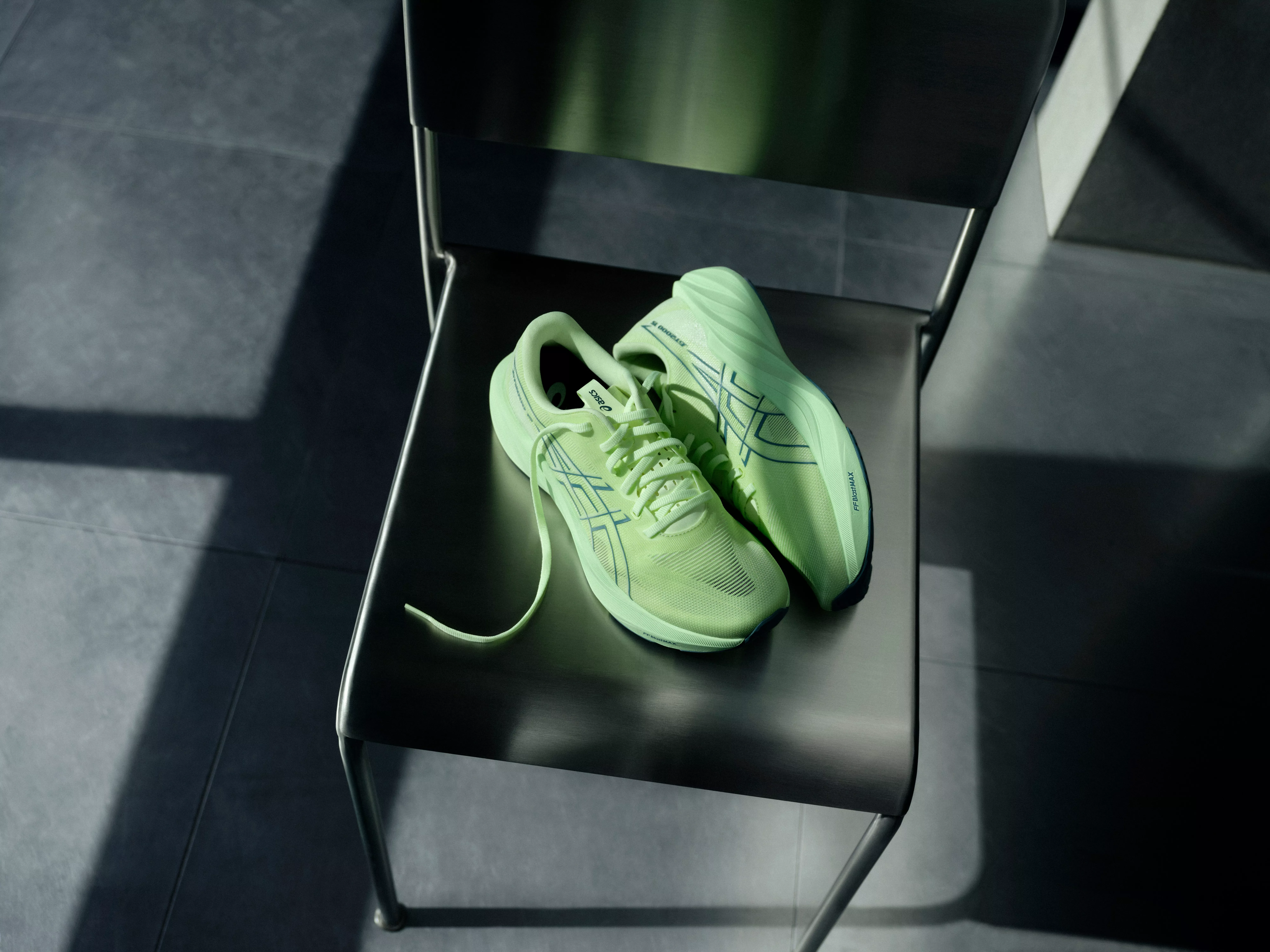 Lime green ASICS running trainers on a metal chair, minimalist indoor product photo with soft shadows
