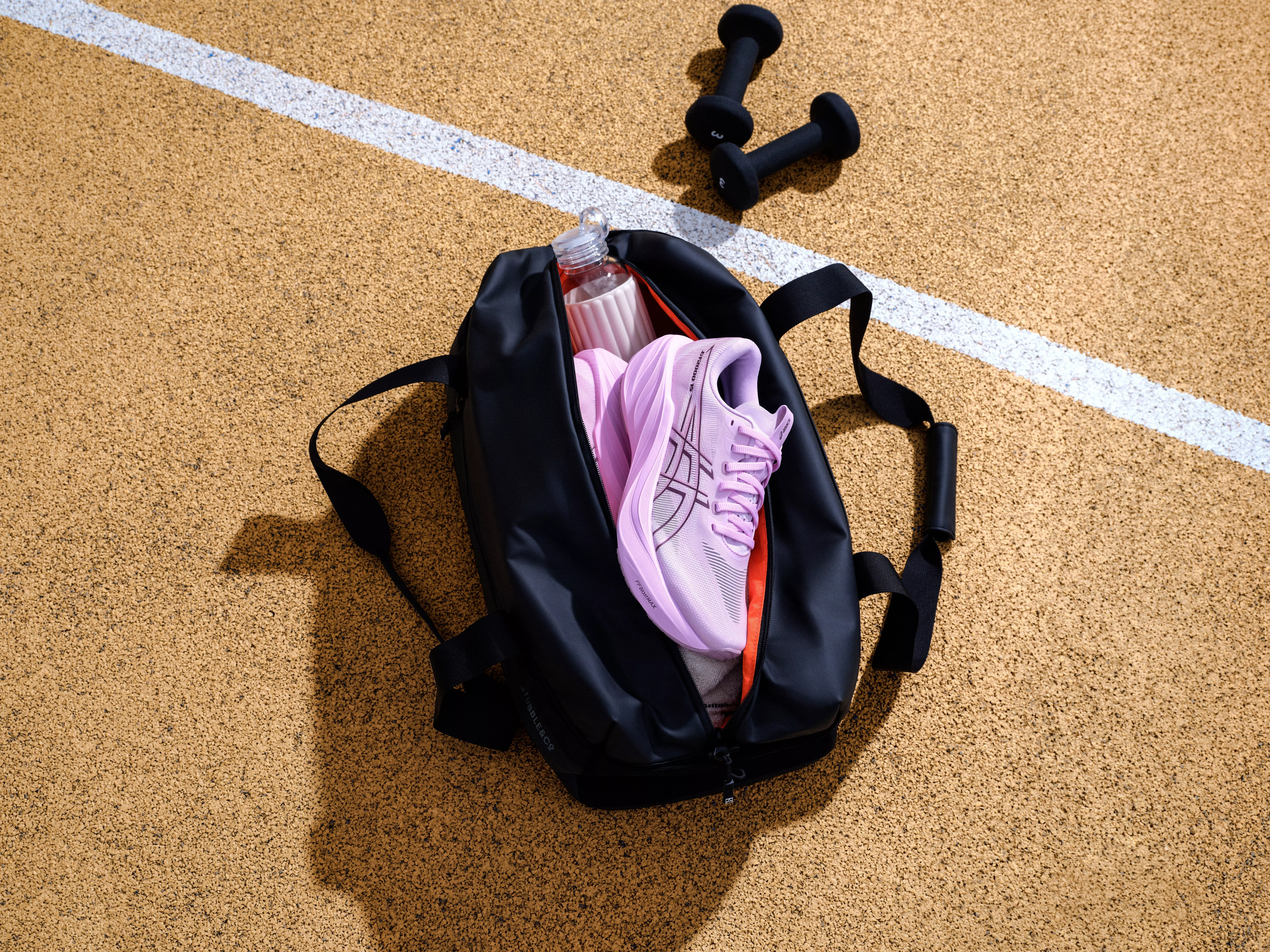 Open gym bag with lilac running shoes and water bottle on track surface, with black dumbbells nearby