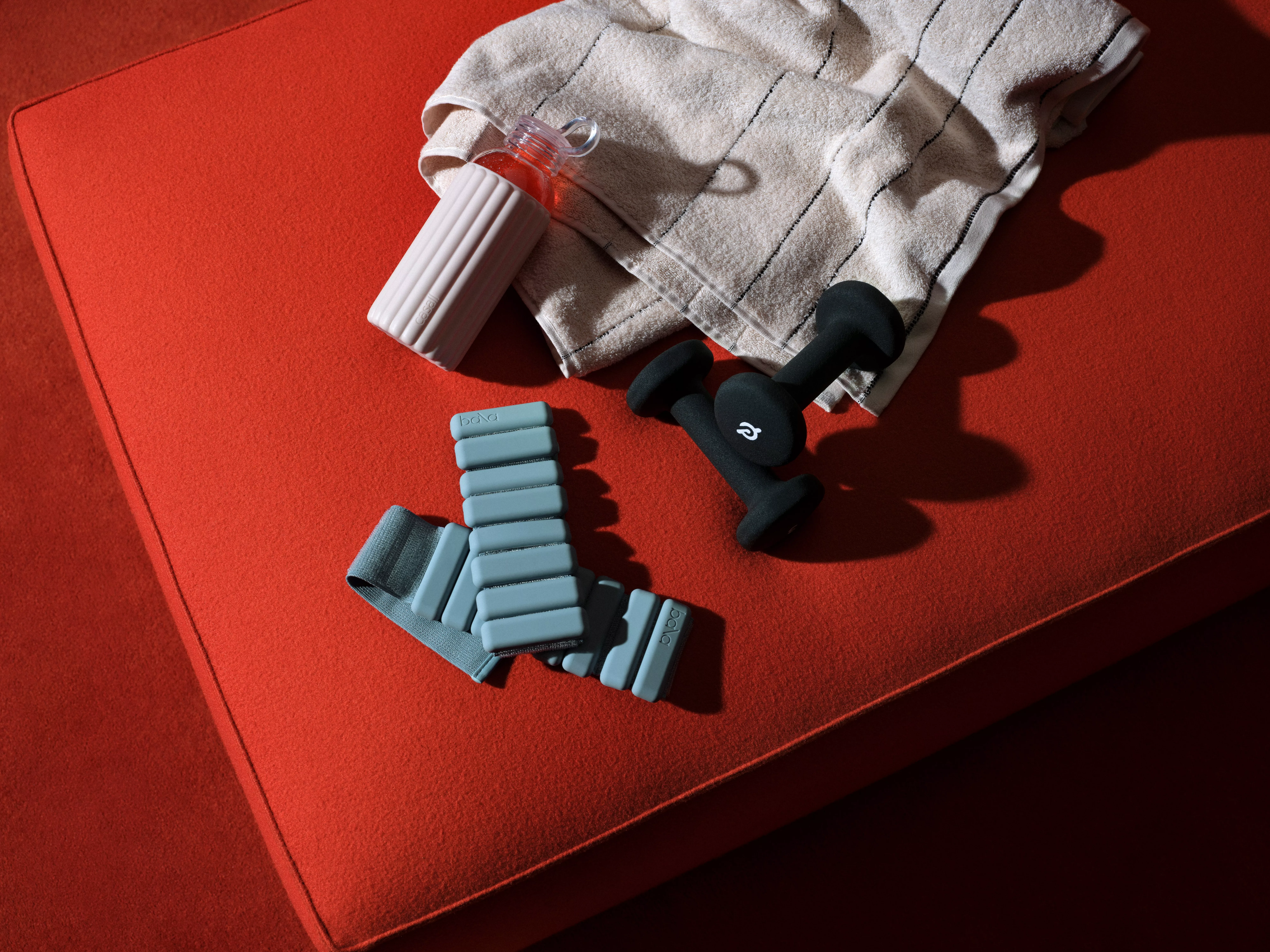 Fitness equipment flat lay on red mat: water bottle, towel, ankle weights and black dumbbells in sunlight