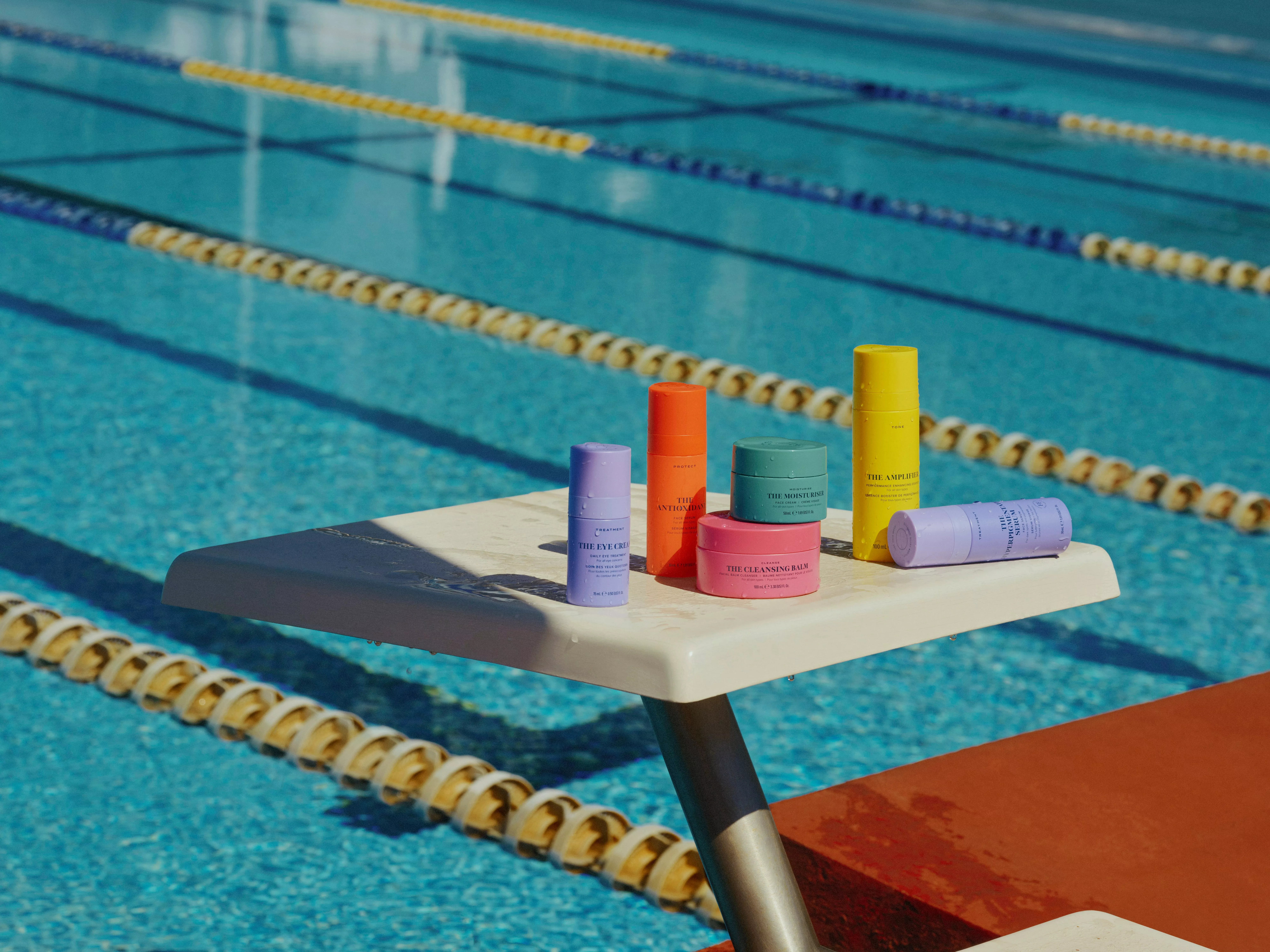 Skincare and sun protection products on a diving board beside an outdoor swimming pool with lane ropes in view