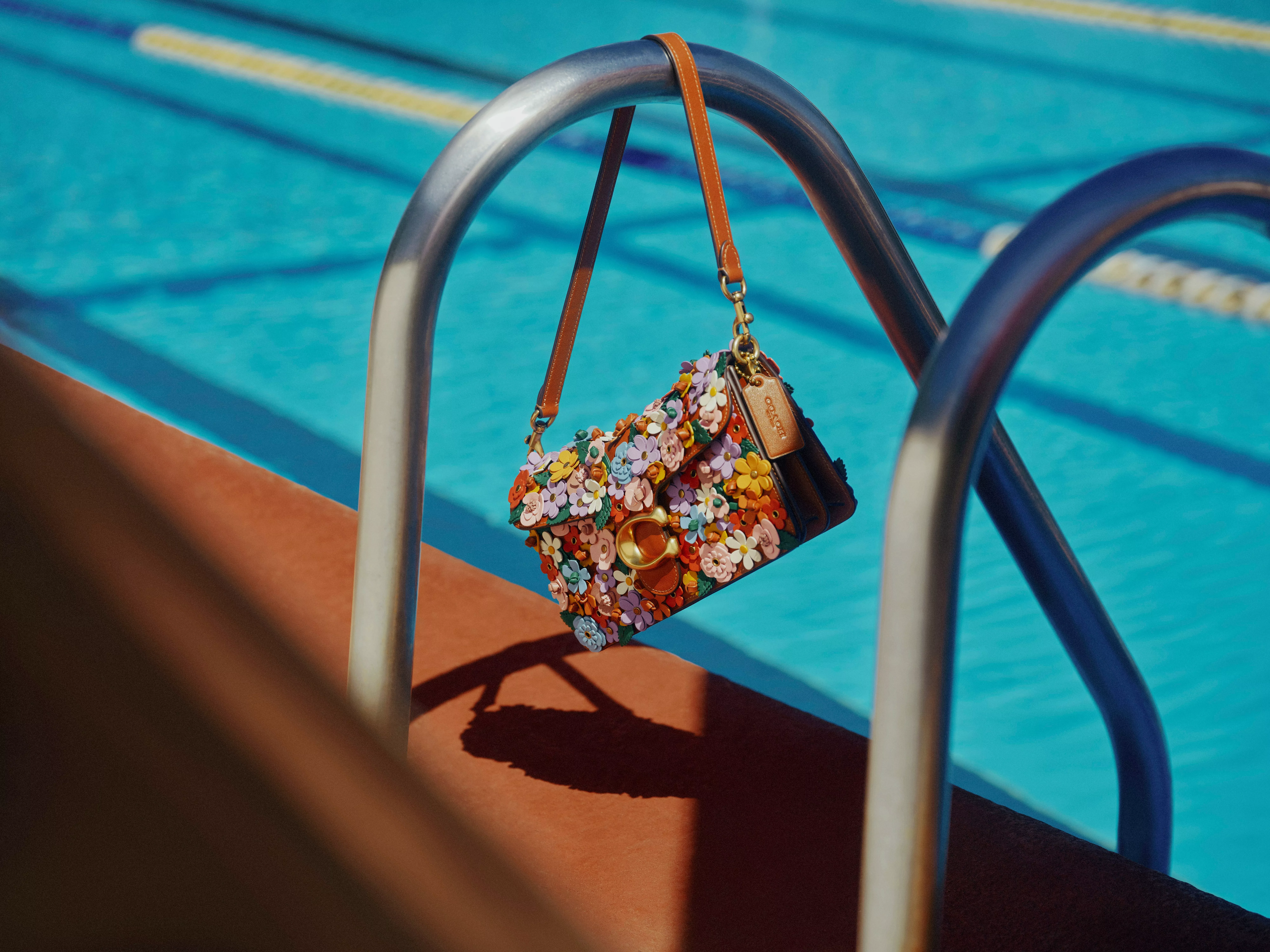 Floral leather shoulder bag hanging on swimming pool ladder, colourful 3D flowers and tan strap in sunlight