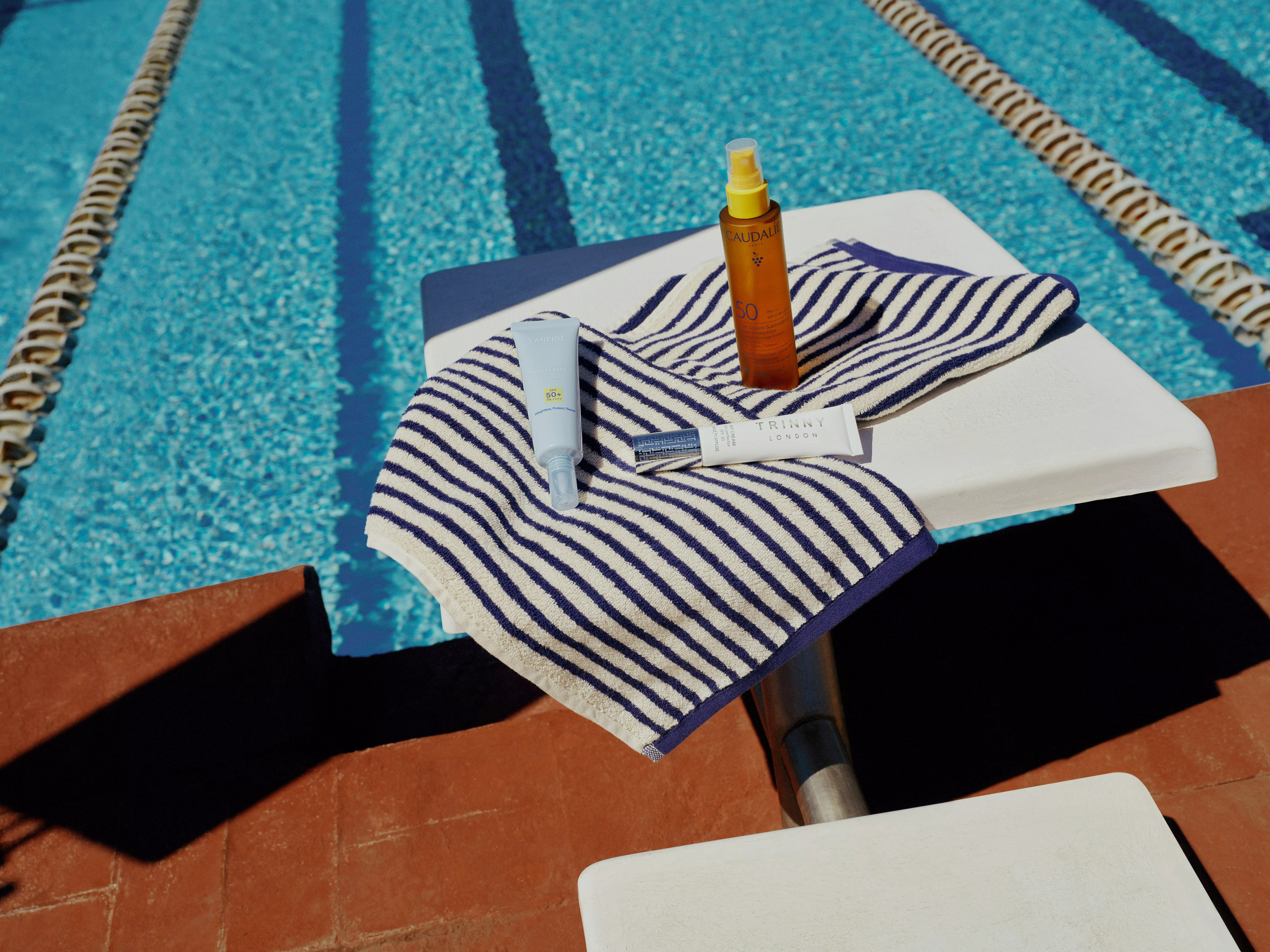 Sun cream and skincare products on a striped towel on a diving board beside an outdoor swimming pool lane lines
