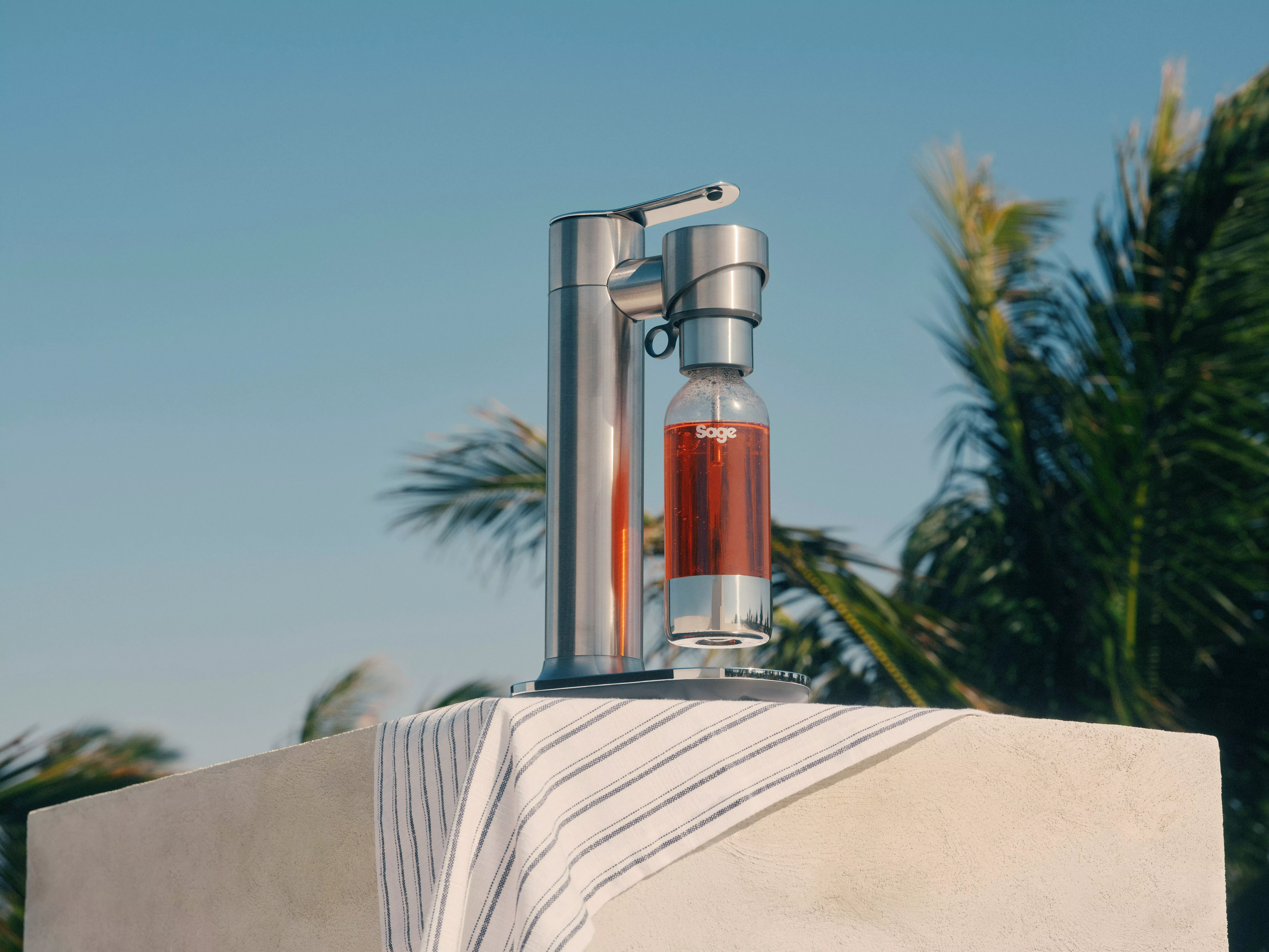 Stainless steel SodaStream-style sparkling water maker with bottle outdoors, blue sky and palm trees backdrop