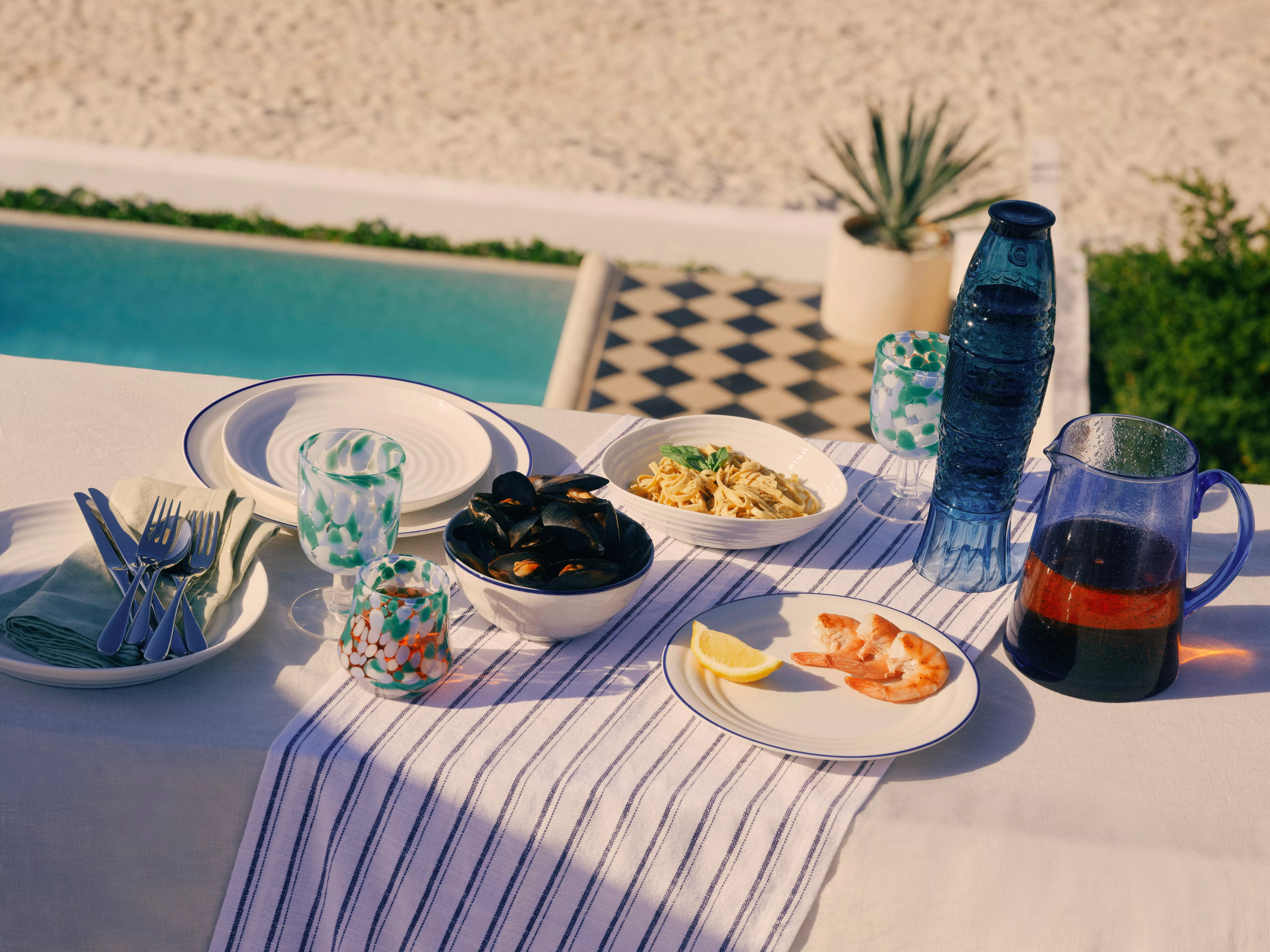 Outdoor poolside table set for seafood lunch with prawns, mussels, pasta, lemon, glassware and water carafe