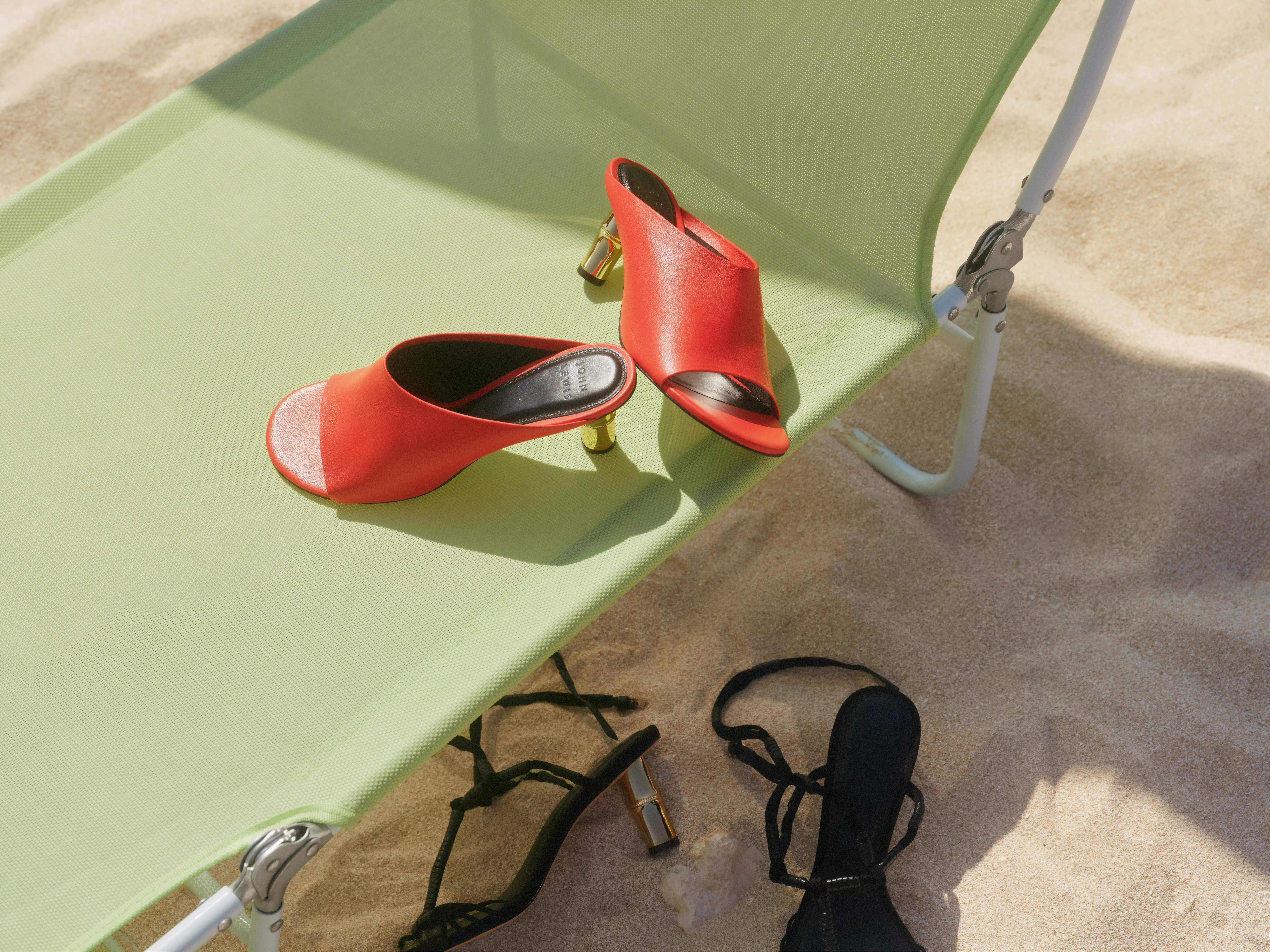 Red heeled mule sandals on green sun lounger, with black heels on sandy beach in bright sunlight