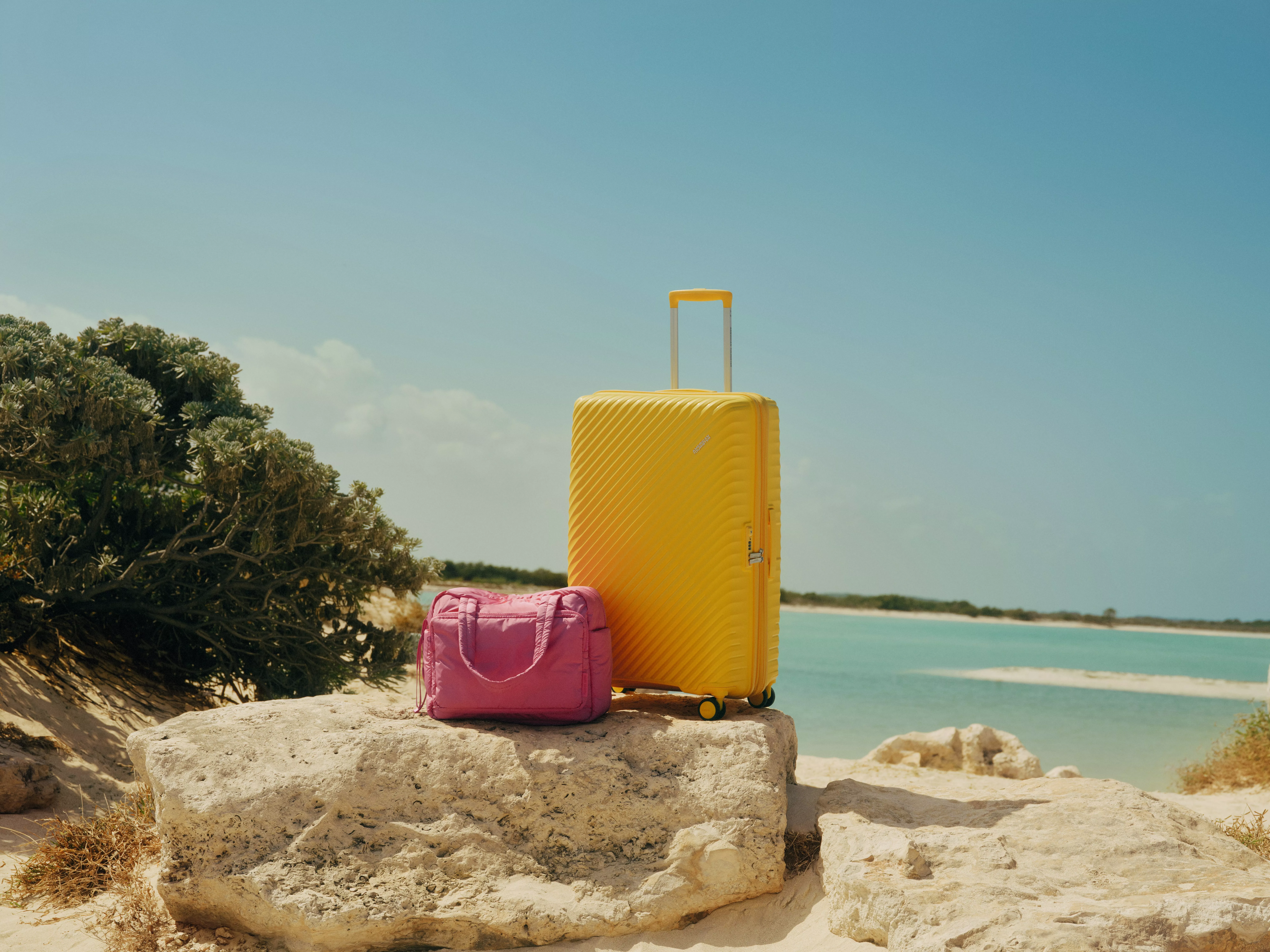 Yellow hard-shell suitcase and pink travel bag on rocks by a turquoise beach, summer holiday luggage scene