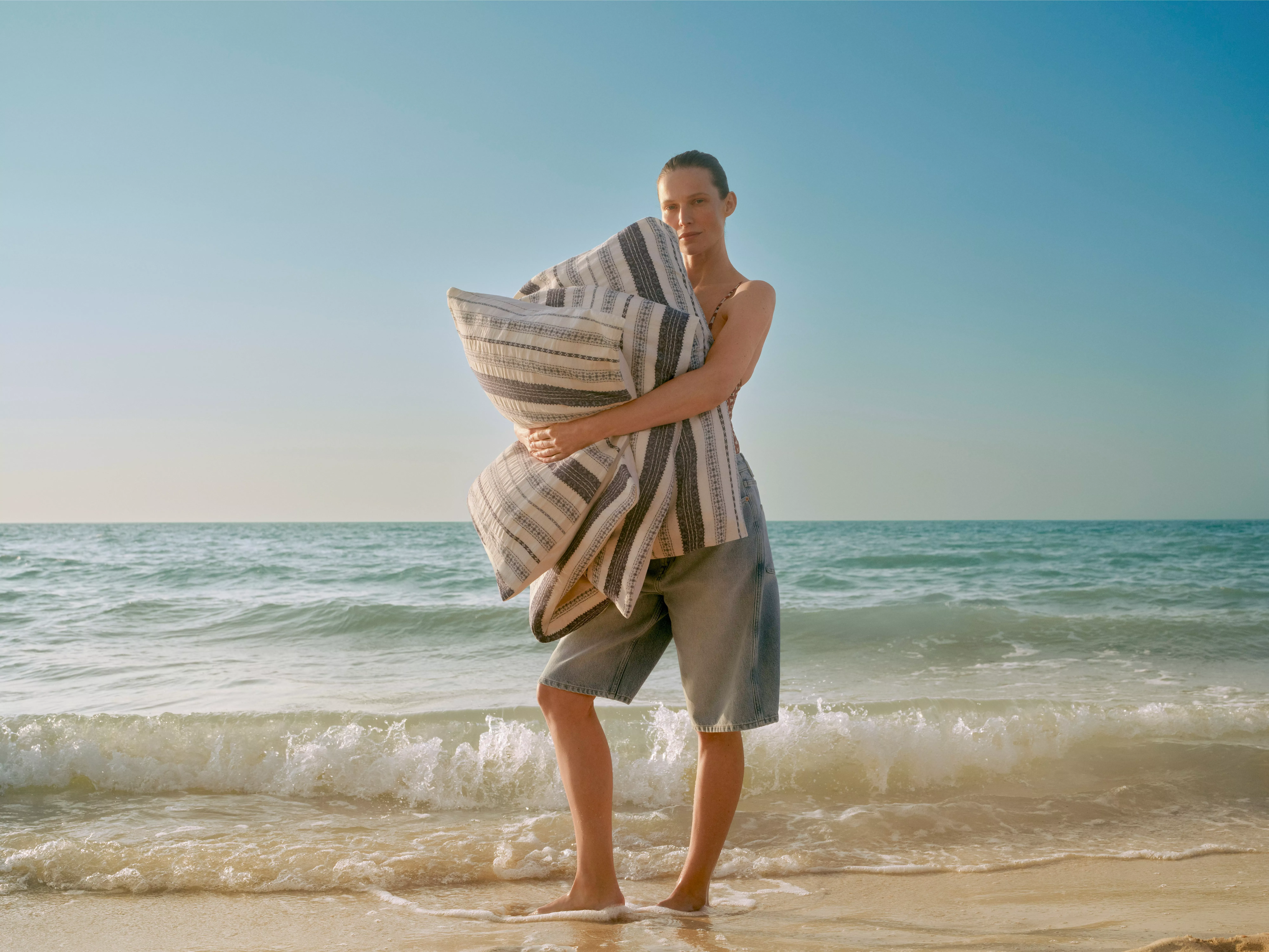 Woman holding striped beach towel on sandy shoreline, gentle waves and clear blue sky in background