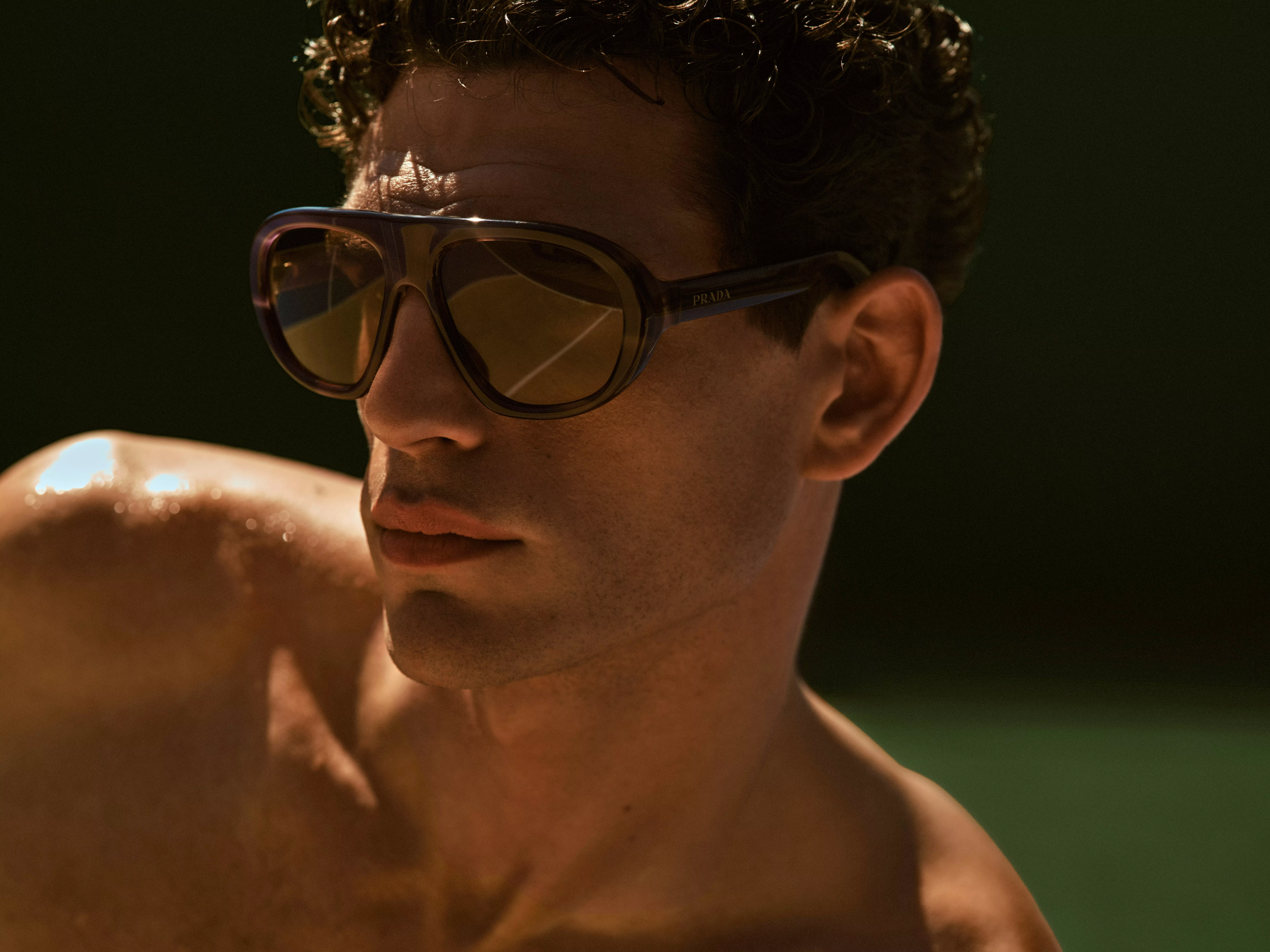 Close-up portrait of a shirtless man outdoors with curly hair and glasses, face blurred in soft light.
