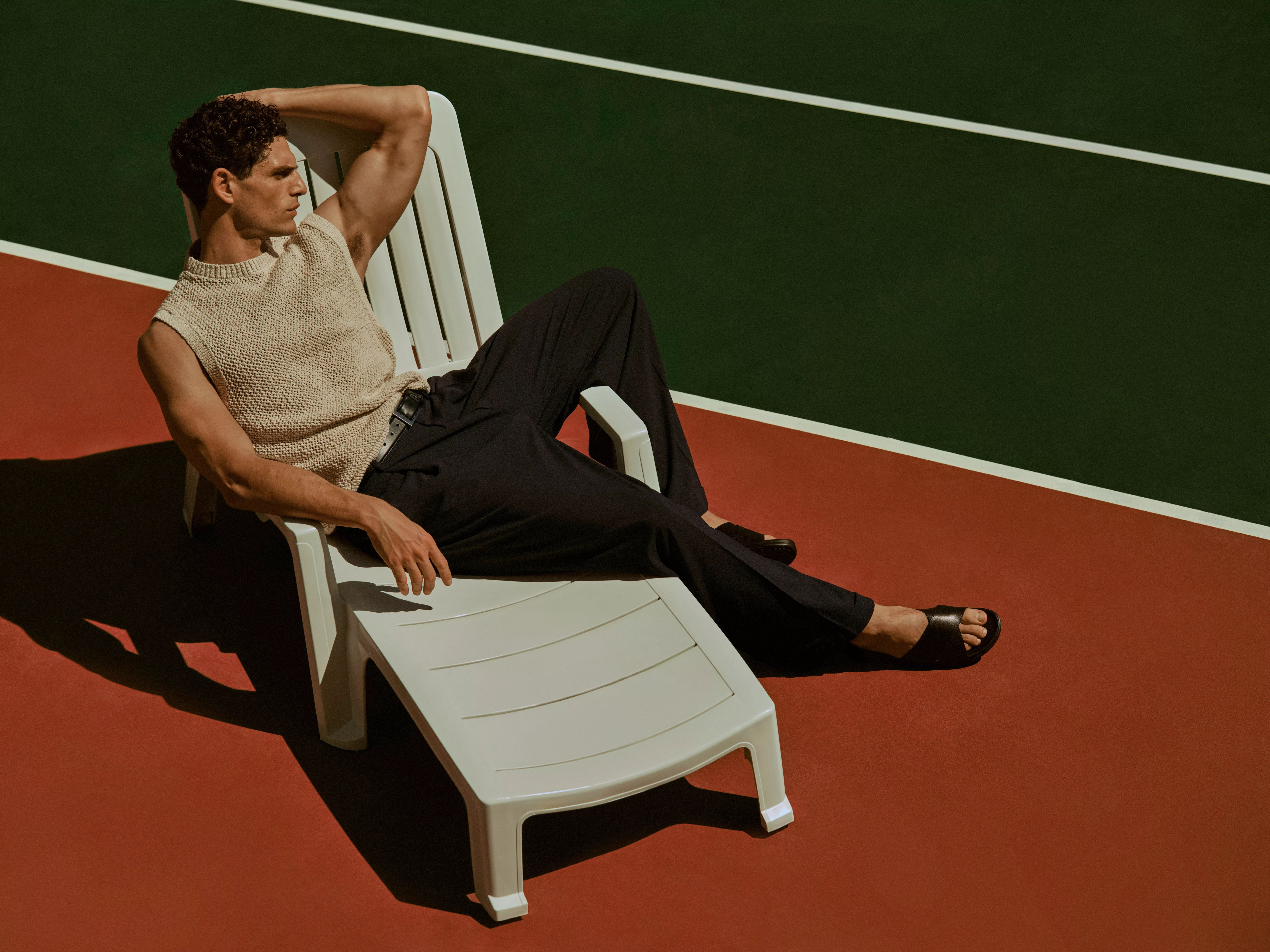 Stylish man reclining on white sun lounger on tennis court, wearing sleeveless knit top, trousers and sandals