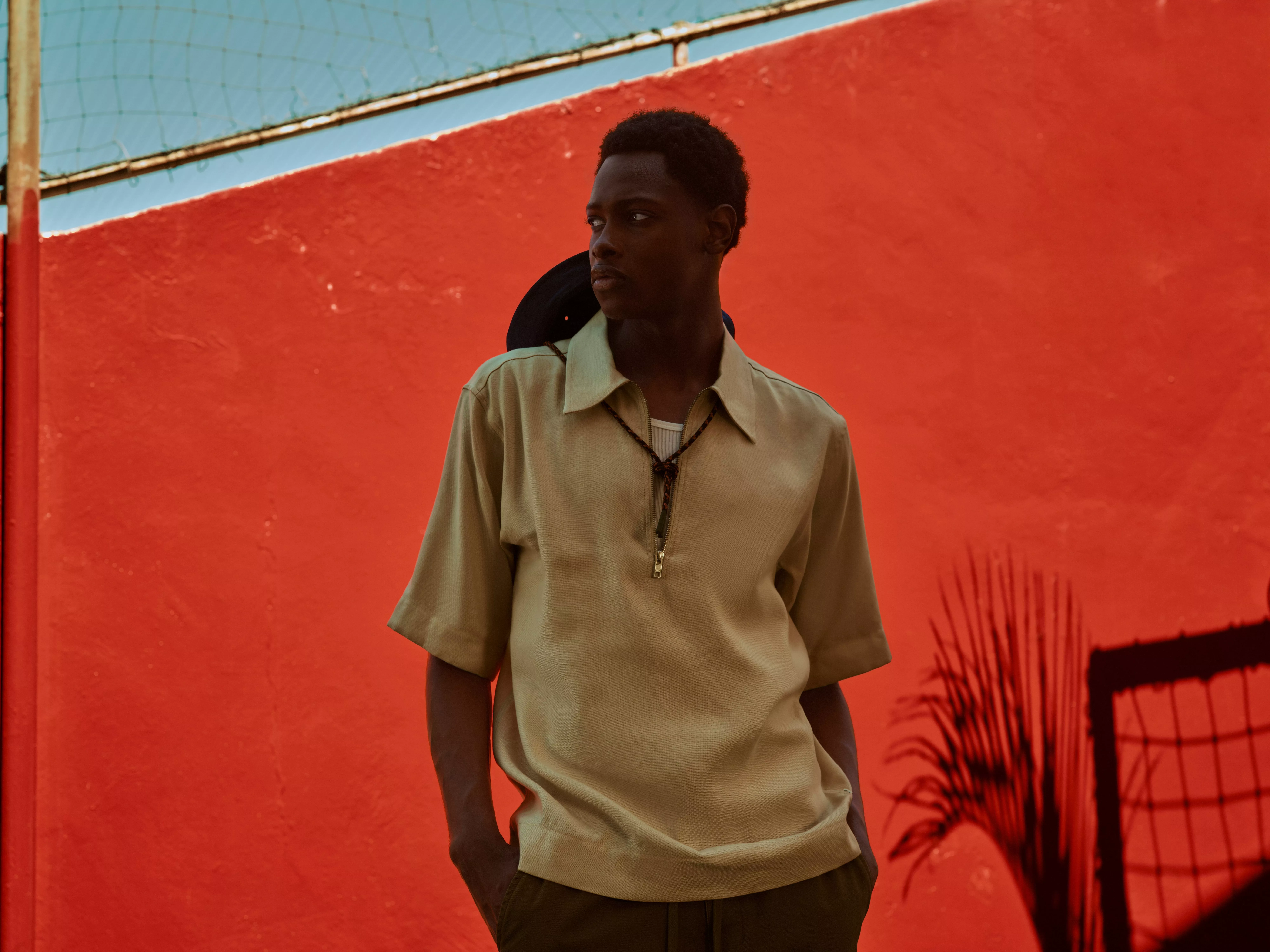 Man wearing a shirt, scarf, shorts and bag leaning on a wall